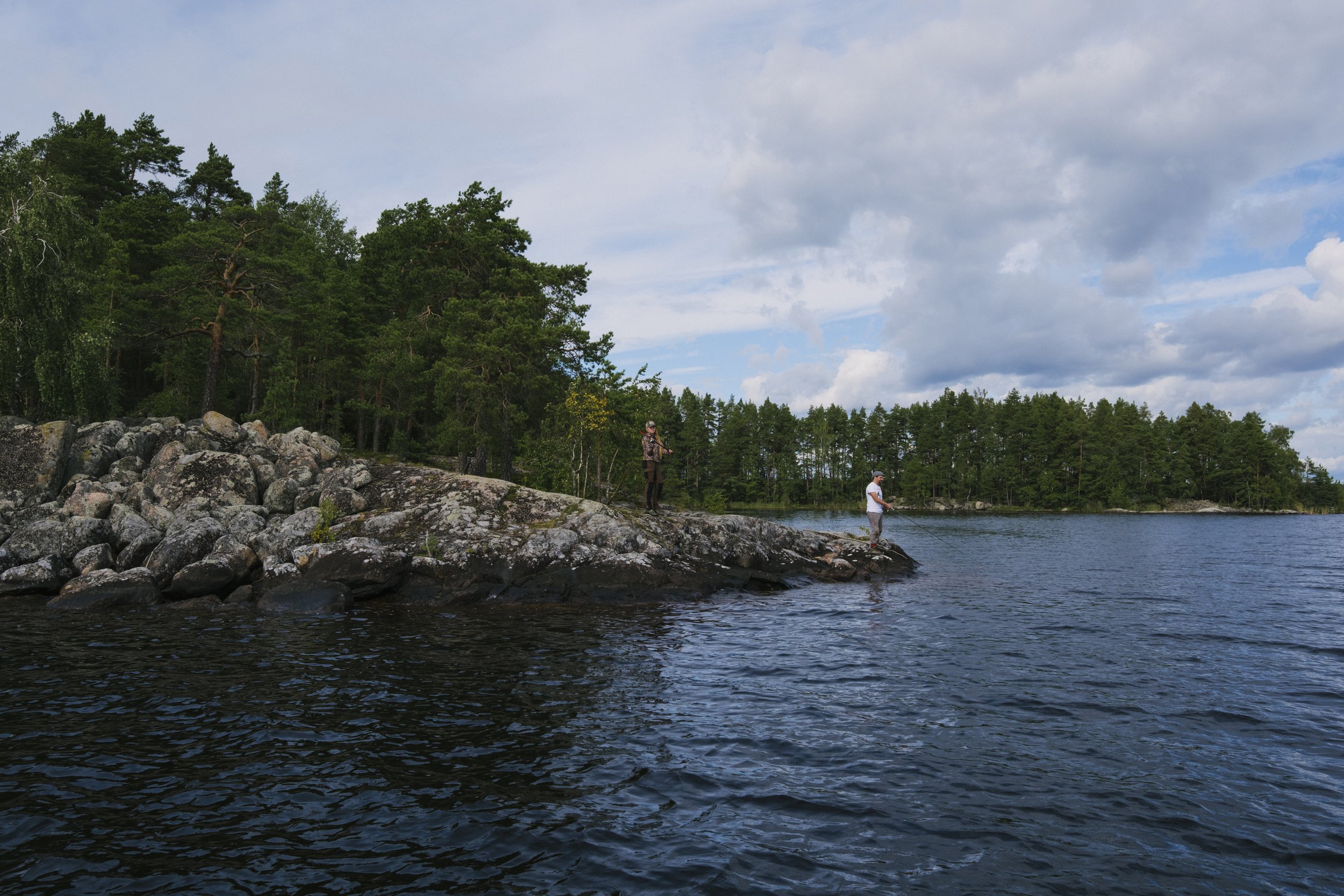 Zwei Menschen beim Angeln am Ufer eines Sees, umgeben von Bäumen und lilafarbenen Blumen, an einem bewölkten Tag.