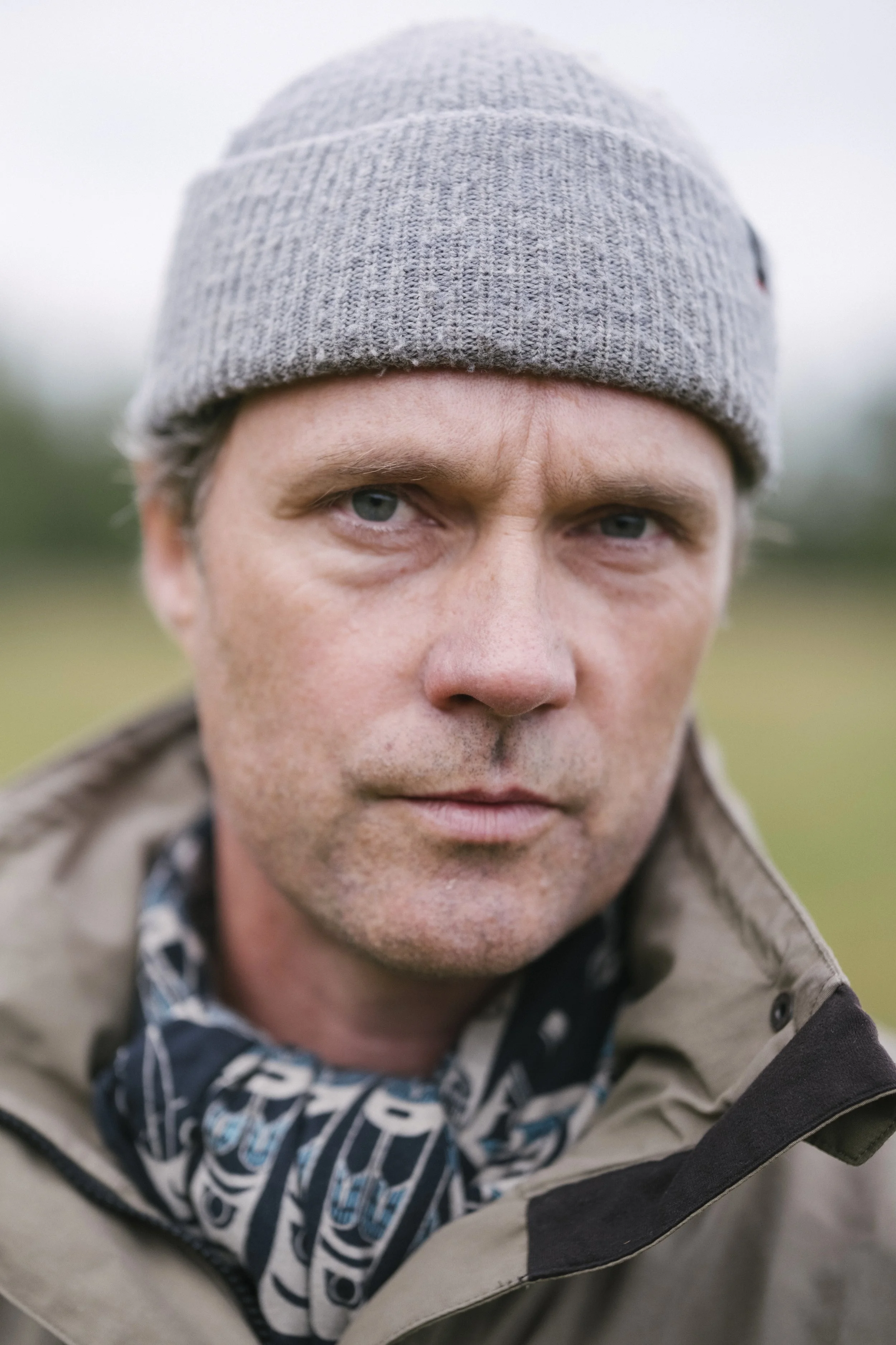 Ein Mann trägt eine graue Mütze und eine Jacke, der Hintergrund ist unscharf und zeigt eine grüne Naturlandschaft.