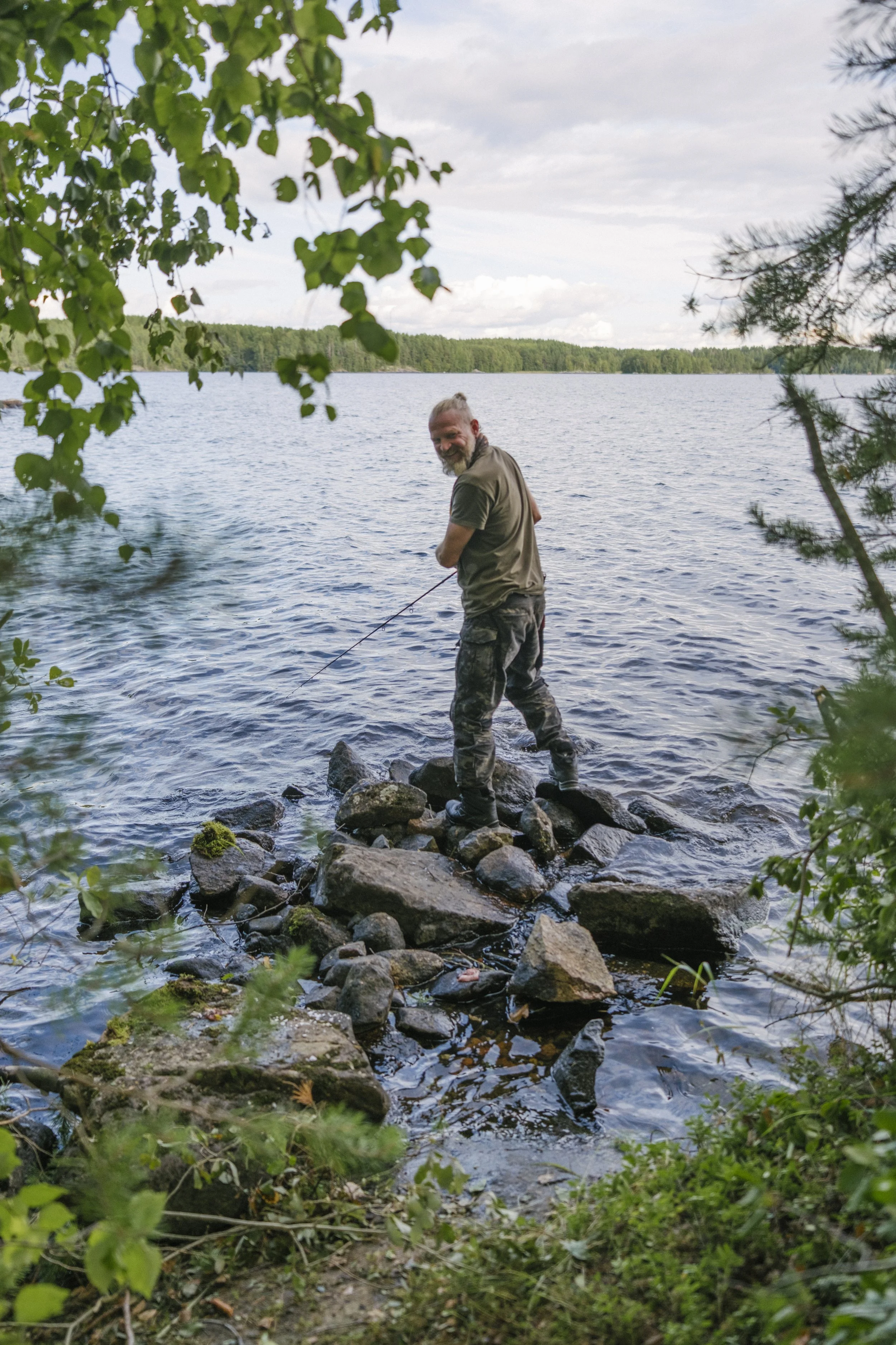 Ein Mann steht am Ufer eines Sees auf Steinen, hält eine Angelrute und lächelt. Der Himmel ist bewölkt, umgeben von Bäumen.
