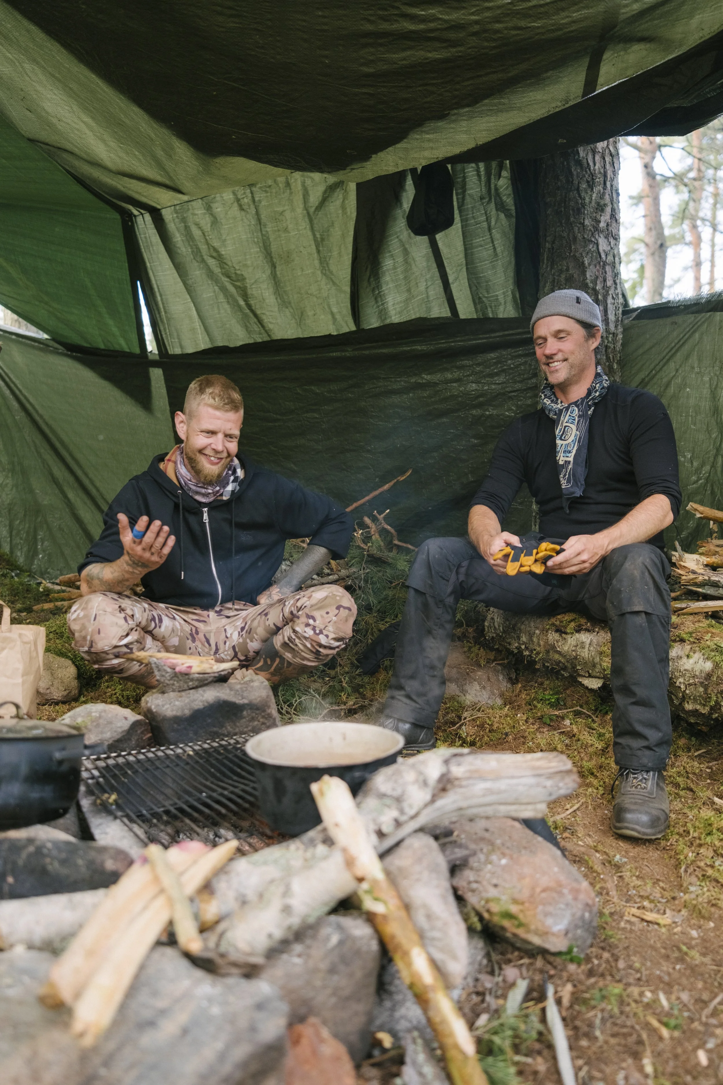 Zwei Männer sitzen in einem grünen Tarnnetz in einem Wald und haben eine Unterhaltung. Sie sind in Outdoor-Bekleidung, umgeben von Feuerstellen mit Steinen, Holz und Kochgeschirr, beim Zelten oder Wildcampen.