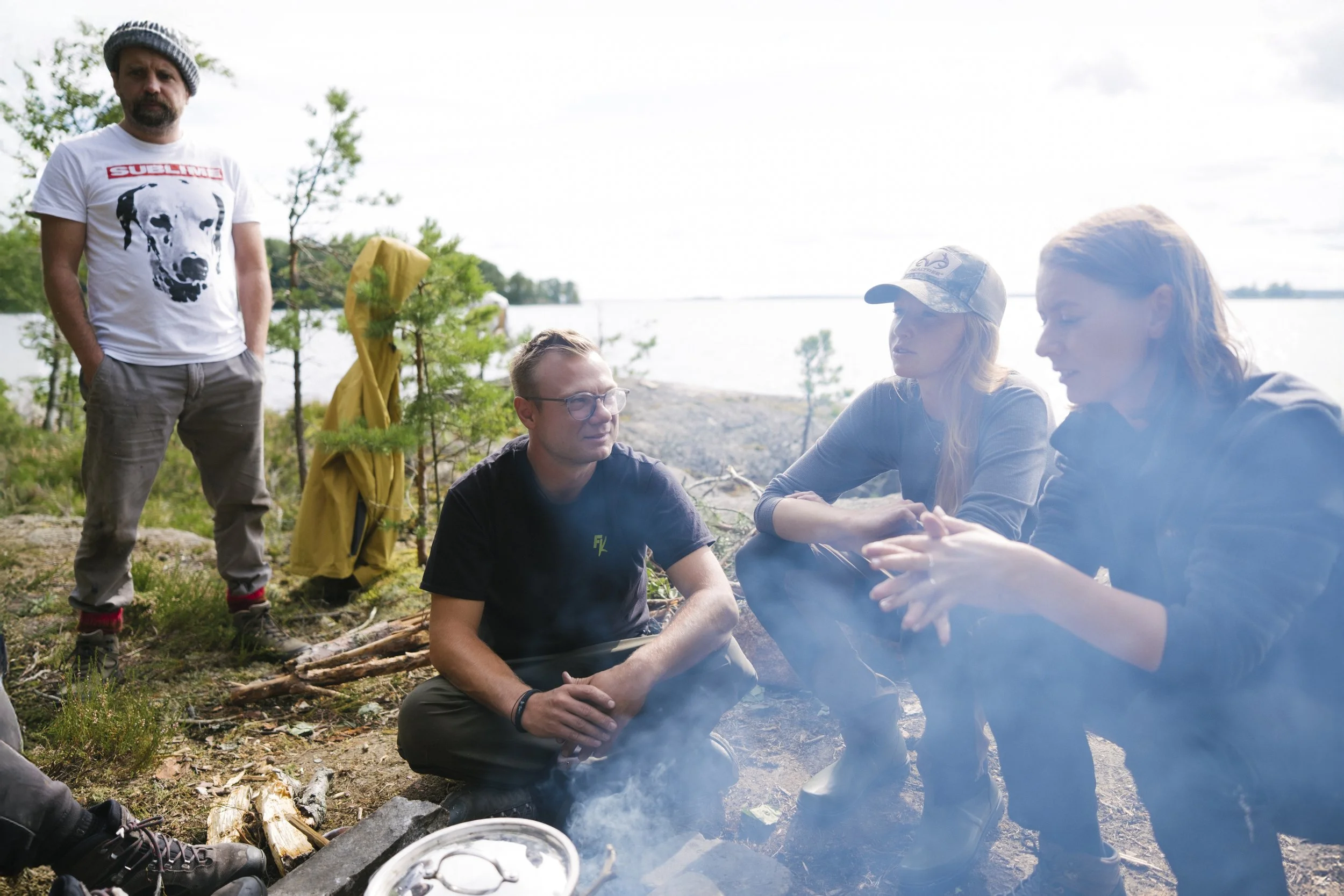 Vier Personen am Lagerfeuer in einer natürlichen Umgebung am Wasser, im Hintergrund Bäume und Wasser, eine Person steht im weißen T-Shirt, die anderen sitzen oder knien rund um das Feuer.