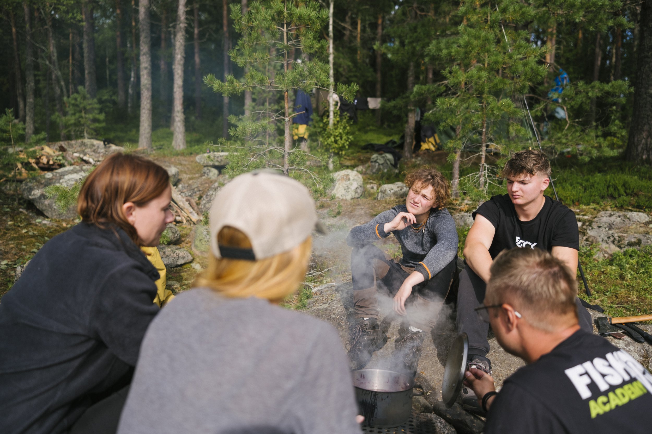 Eine Gruppe junger Menschen sitzt rund um ein Lagerfeuer in einem Wald. Einige schauen in den Topf, während andere sich unterhalten.