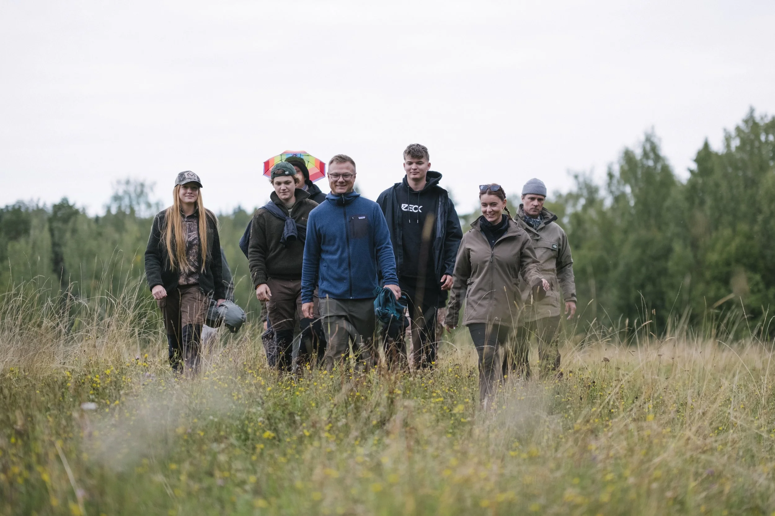 Eine Gruppe von sieben Menschen wandert durch eine grüne Wiesenlandschaft bei bewölktem Himmel, einige tragen Jacken und Hüte, und eine Person hält einen bunten Regenschirm.