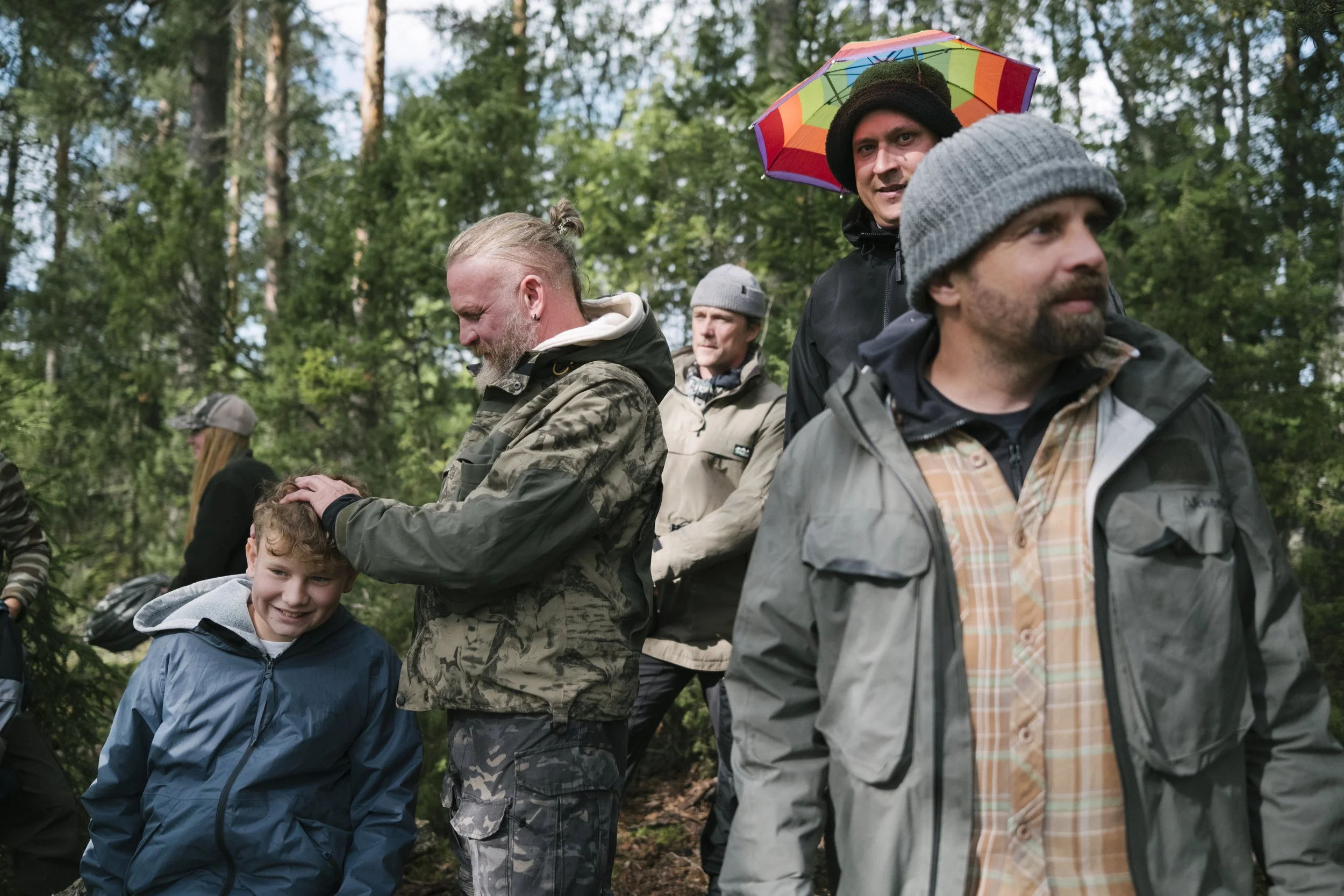 Gruppe von Menschen in einem Wald, einige lachen und schauen entspannt, ein Mann hält einen kleinen Jungen süß und streichelt seinen Kopf, während ein anderer Mann mit Regenschirm im Hintergrund steht.