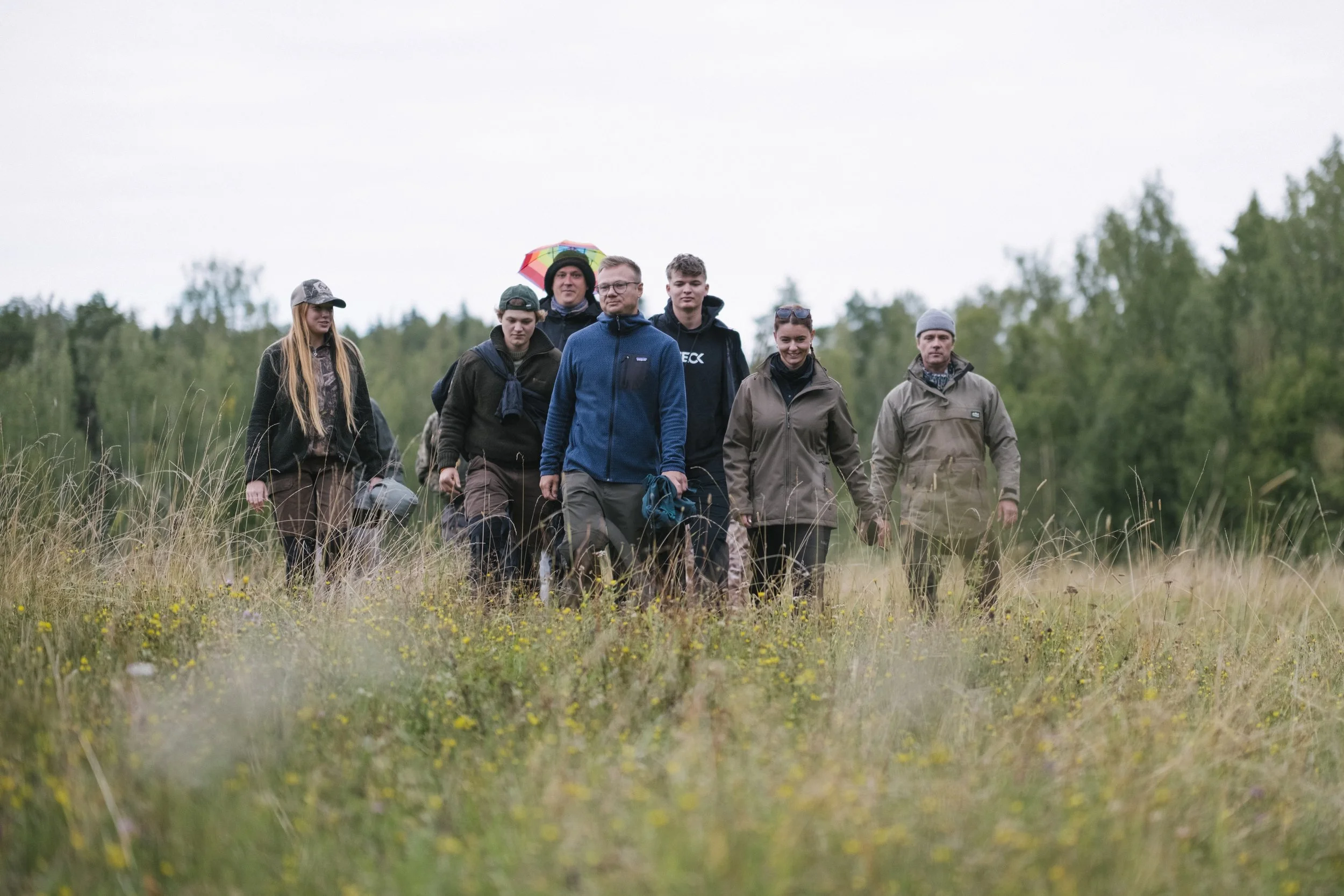 Eine Gruppe von acht Menschen wandert durch ein waldiges und grasiges Gebiet bei bewölktem Himmel, einige tragen Jacken und Mützen, eine Person hält einen bunten Regenschirm.