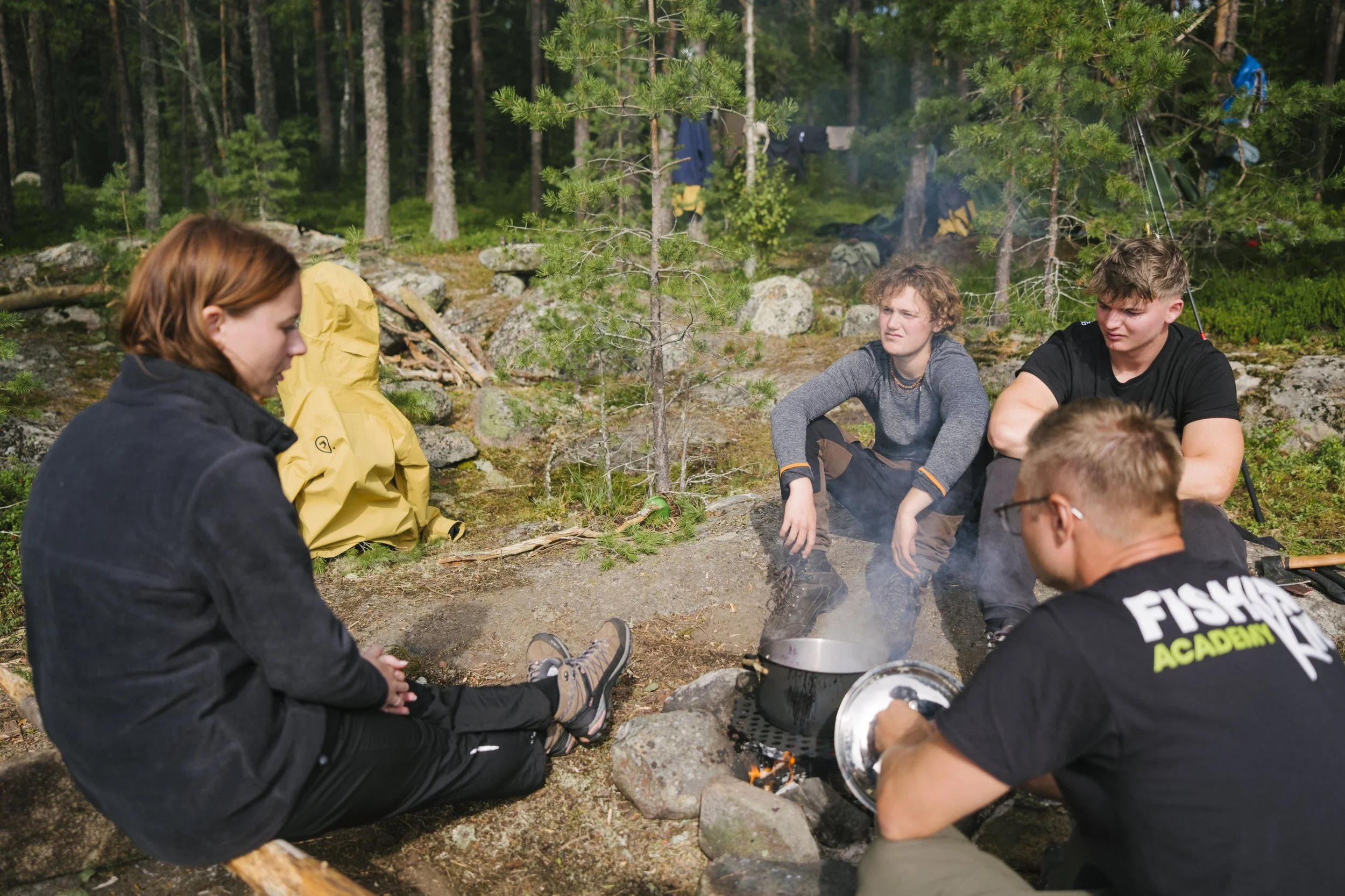 Eine Gruppe Jugendlicher sitzt um ein Feuer im Wald, einige schauen skeptisch, während einer mit einer Pfanne beschäftigt ist.