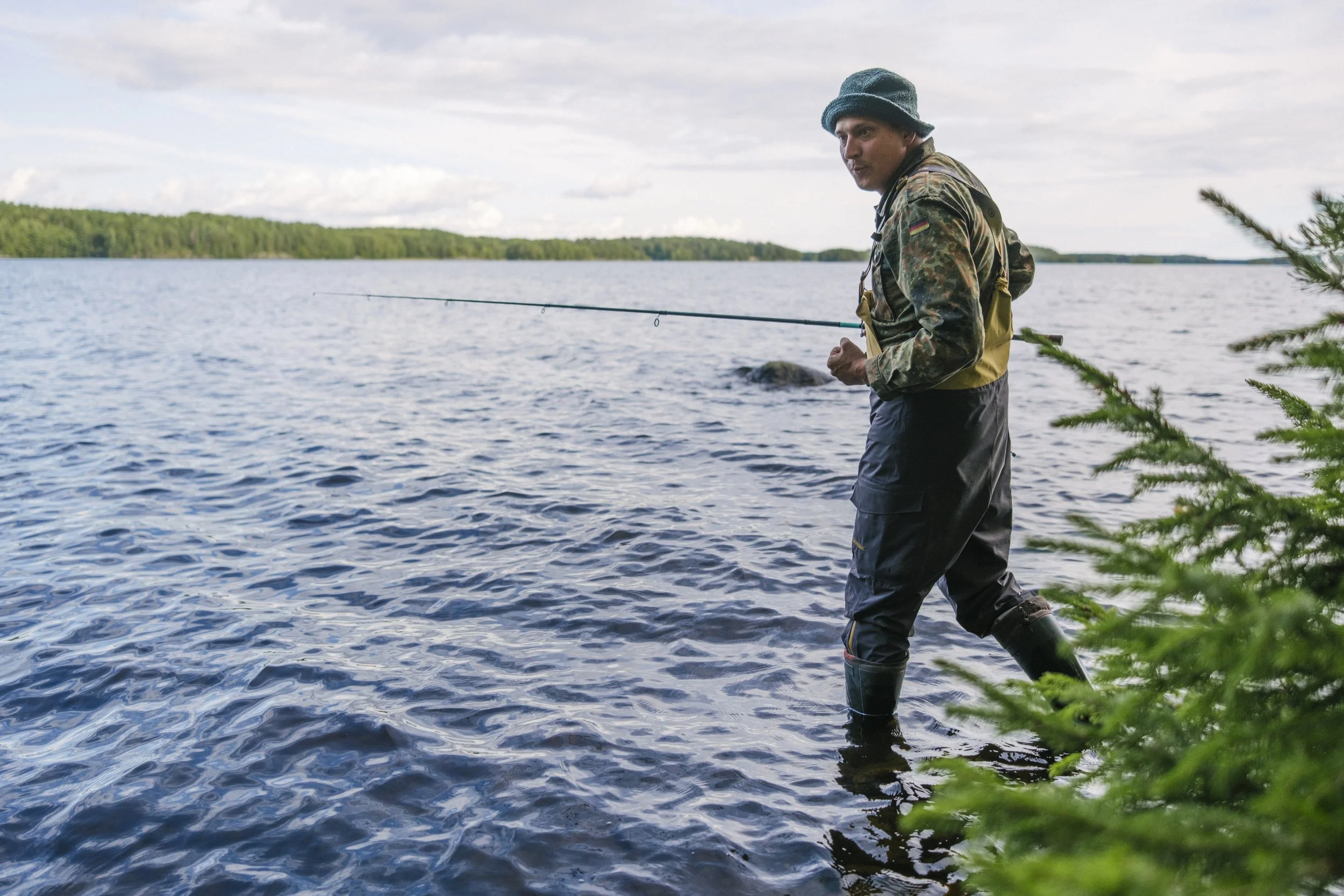 Ein Mann beim Angeln im Wasser, trägt eine Jacke, Gummistiefel und einen Hut, im Hintergrund ein See und bewachsene Ufer.