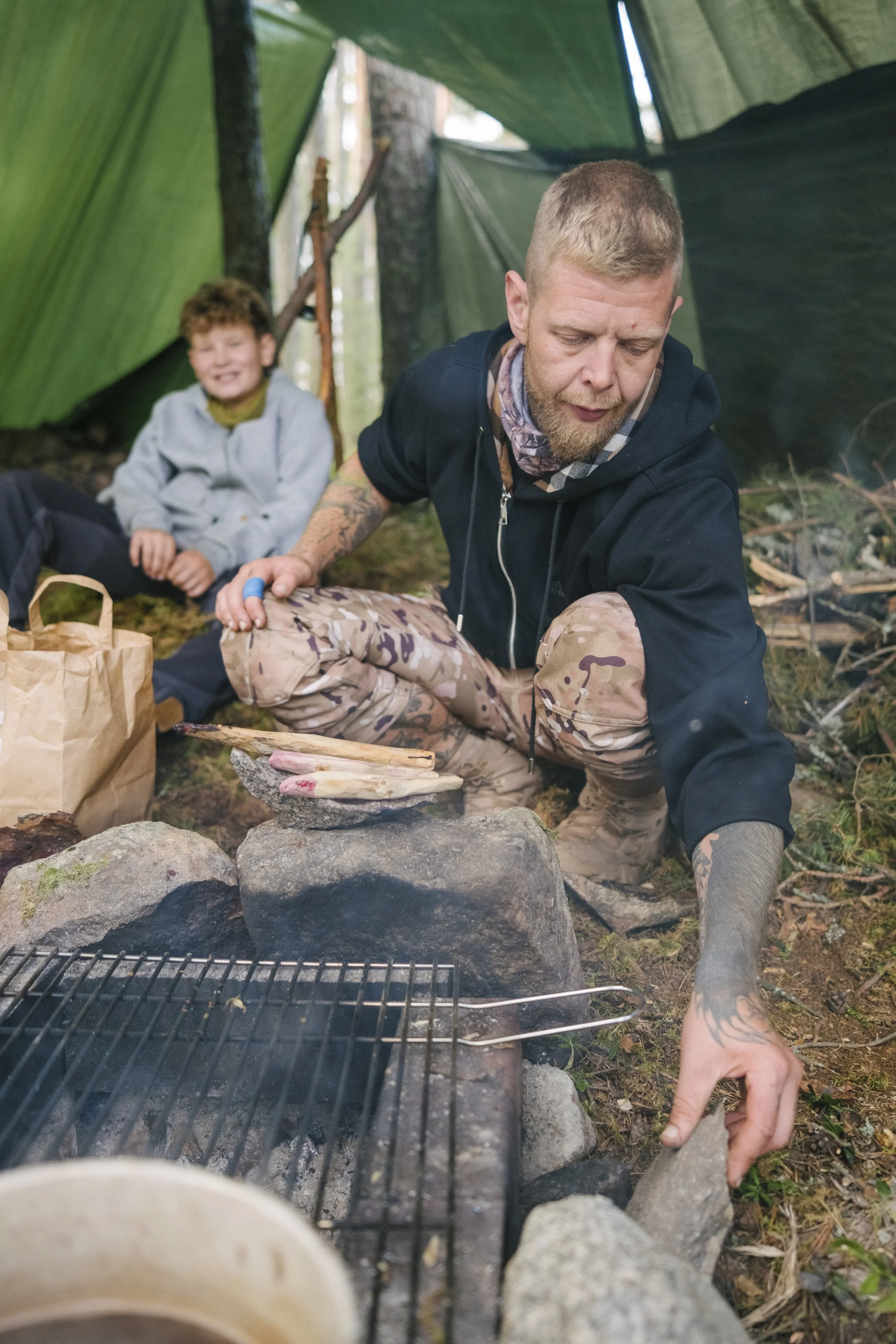 Ein Mann kocht beim Zelten im Wald, im Hintergrund sitzt ein Junge auf dem Boden und lächelt.