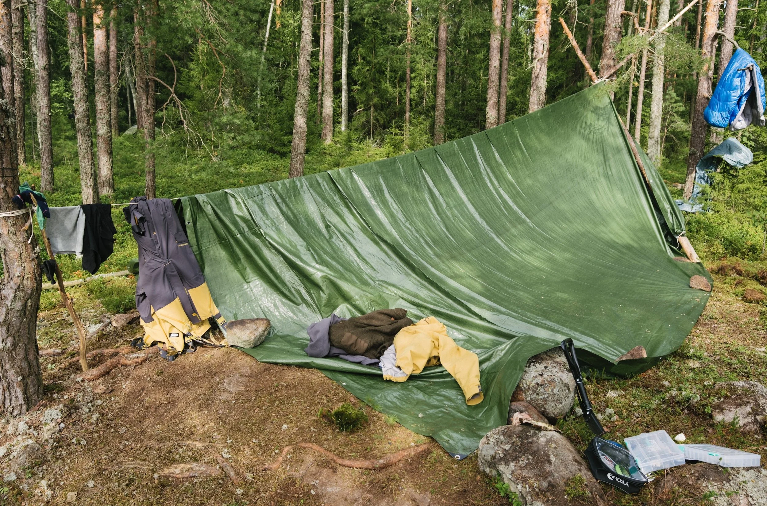Ein provisorisches Zelt aus grüner Folie in einem Wald, mit Kleidung und Ausrüstung drumherum.