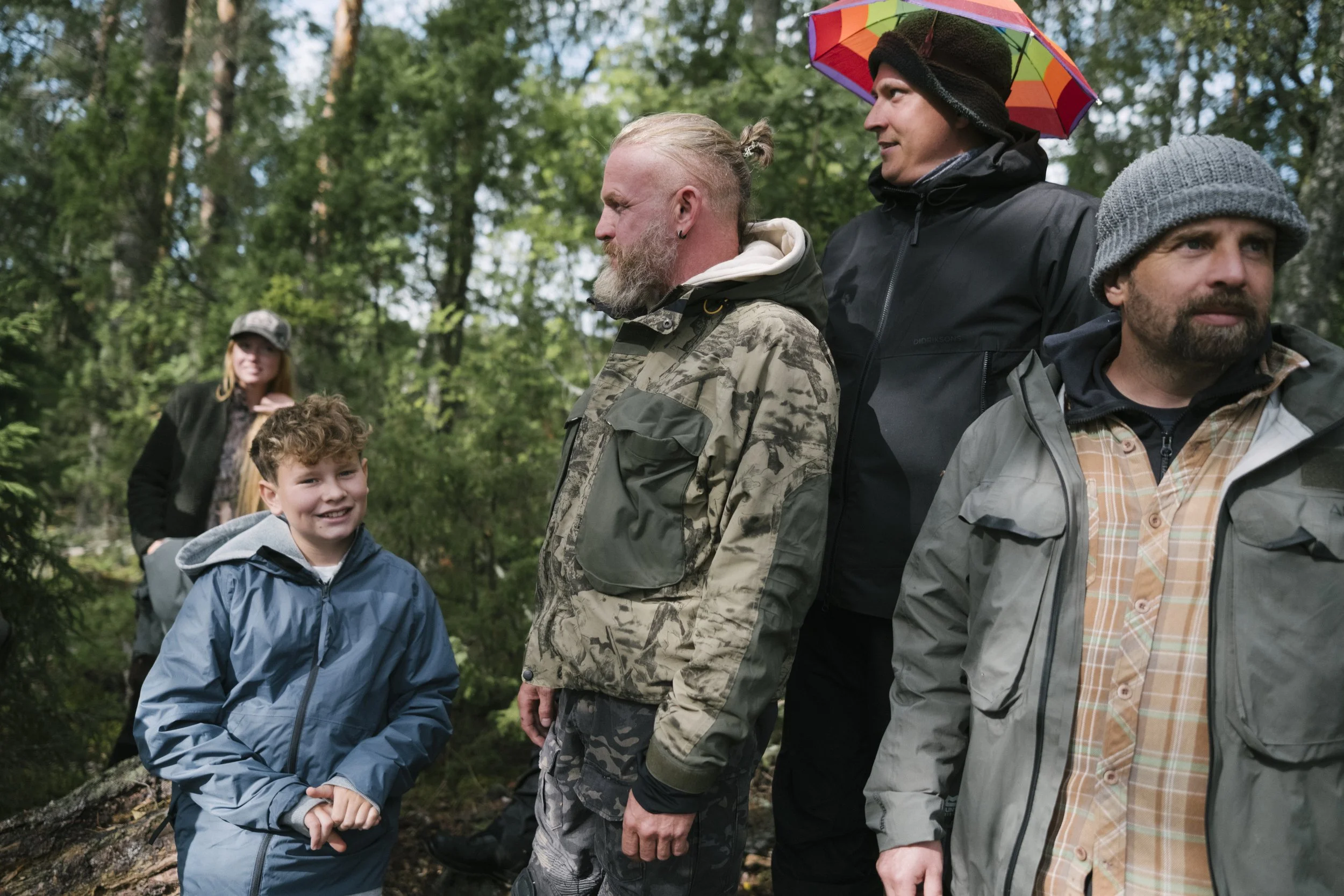 Gruppe von Menschen in einem Wald, einige tragen Jacken und Mützen, eine Person hält einen bunten Regenschirm.