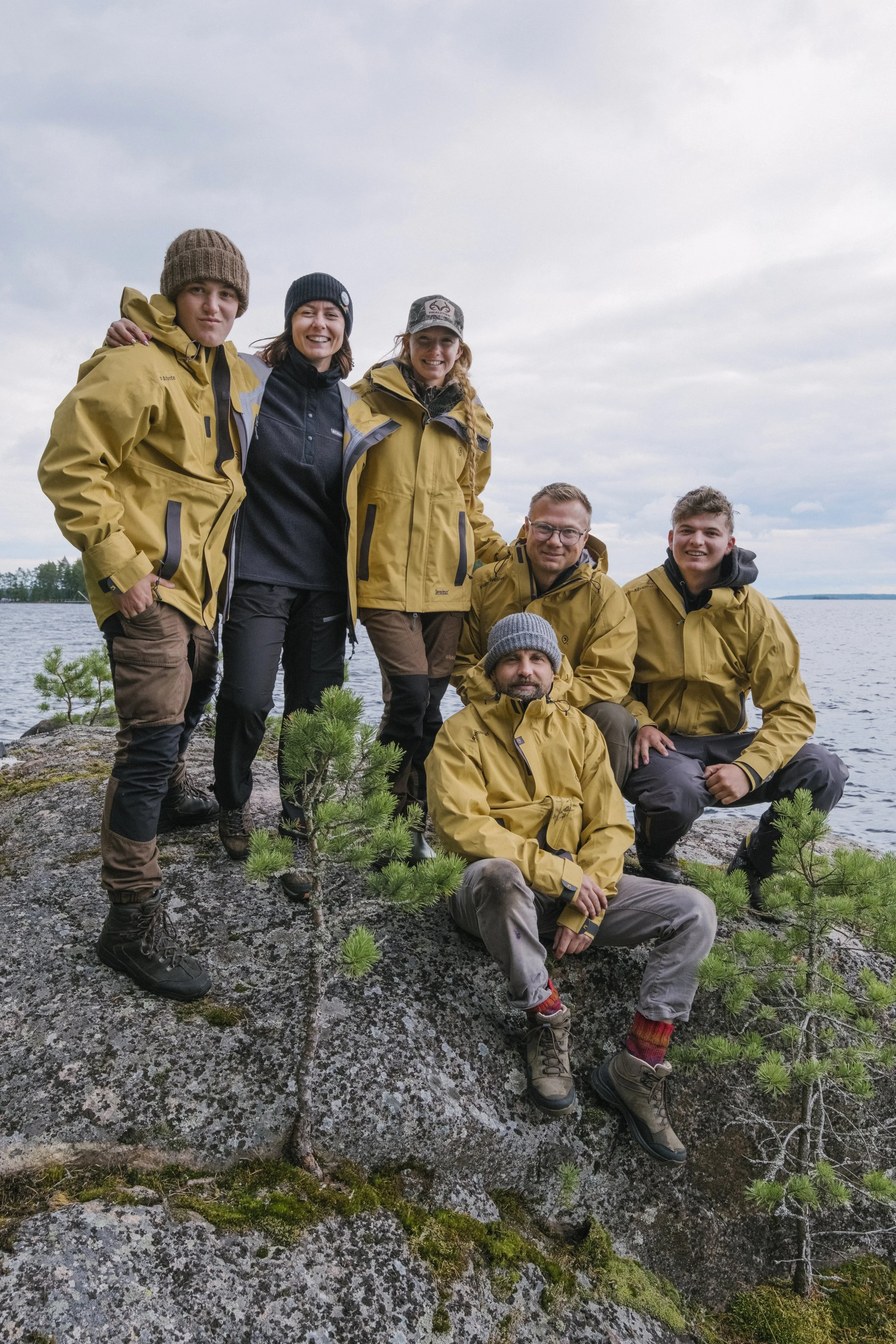 Eine Gruppe von sechs Personen in gelben Outdoor-Jacken steht und sitzt auf einem felsigen Ufer am See mit einem bewölkten Himmel im Hintergrund.