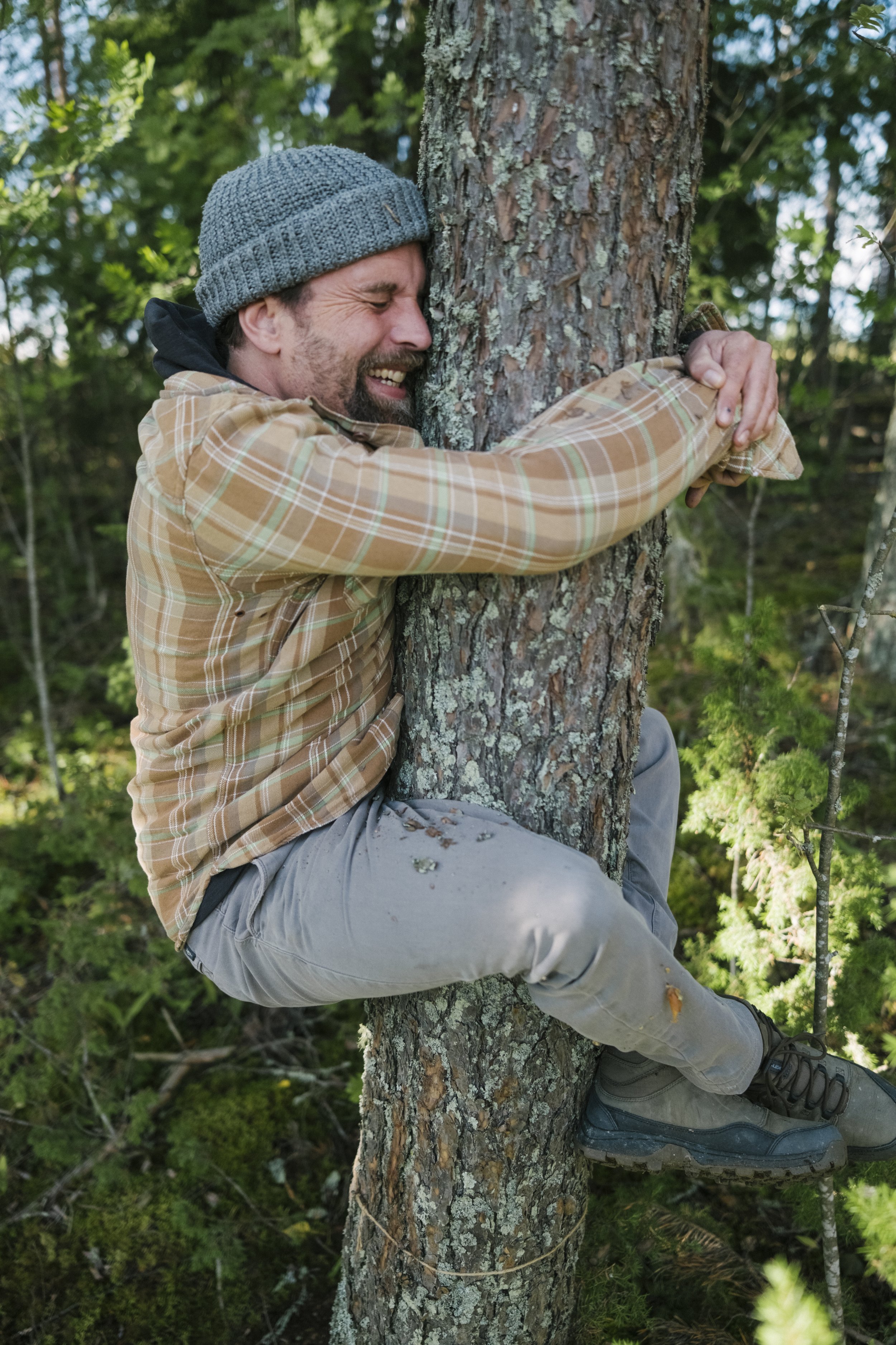 Ein lachender Mann in einem karierten Hemd und grauem Mütze umarmt einen Baum im Wald.