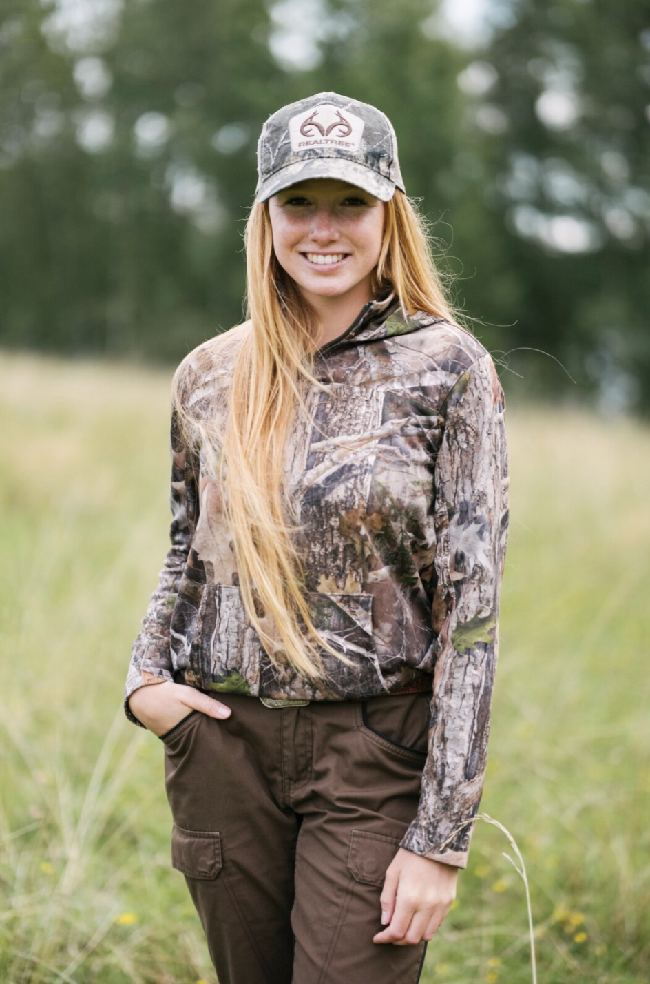 Junge Frau in Camouflage-Kleidung und Camouflage-Cap im Freien, vermutlich beim Wandern oder Jagen, mit grüner Wiese und Bäumen im Hintergrund.