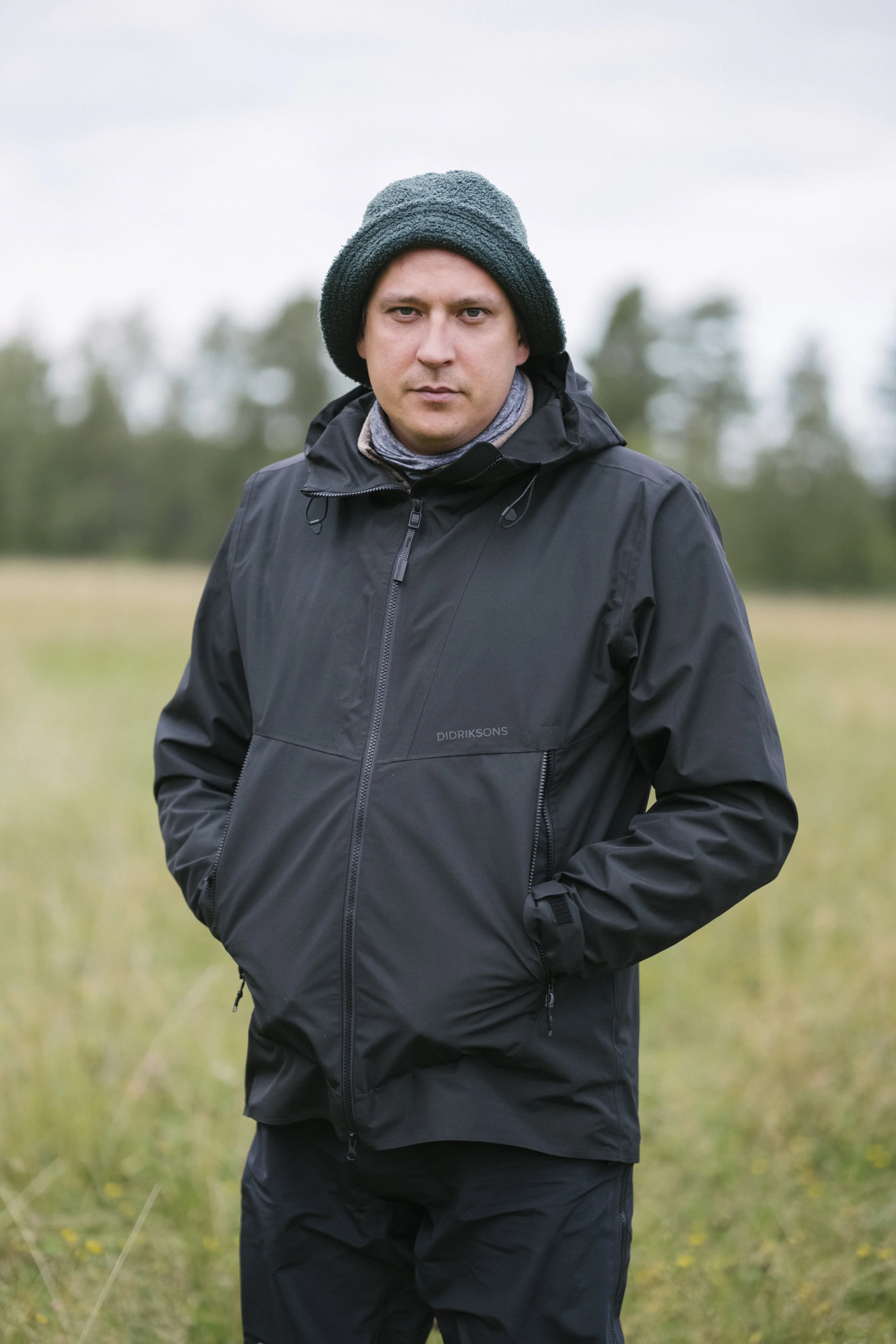 Mann steht in der Natur bei bewölktem Wetter, trägt schwarze Jacke und Mütze, blickt ernst in die Kamera.