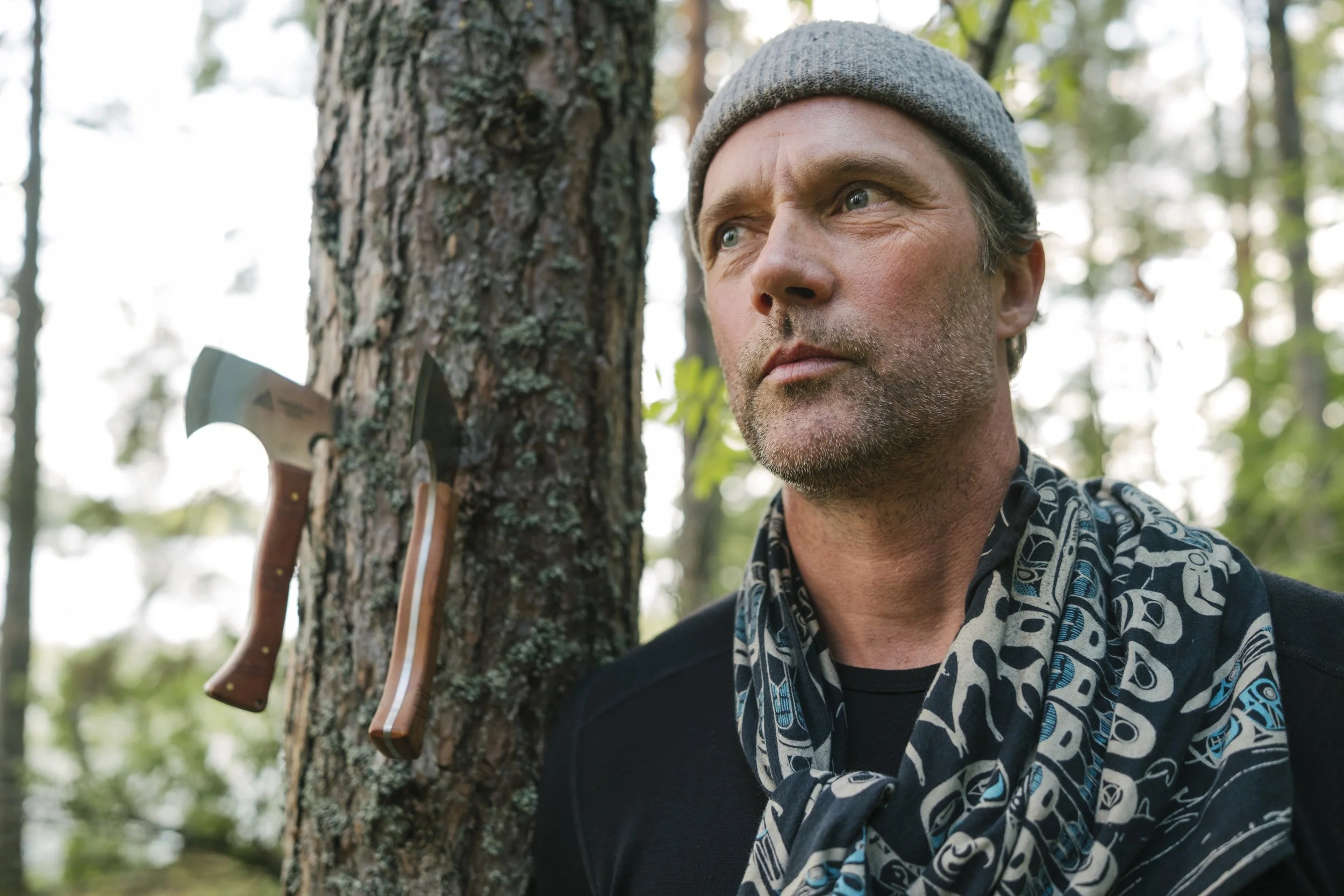 Ein Mann steht an einem Baum im Wald, neben dem ein Axt und ein Messer an den Baum gehängt sind.