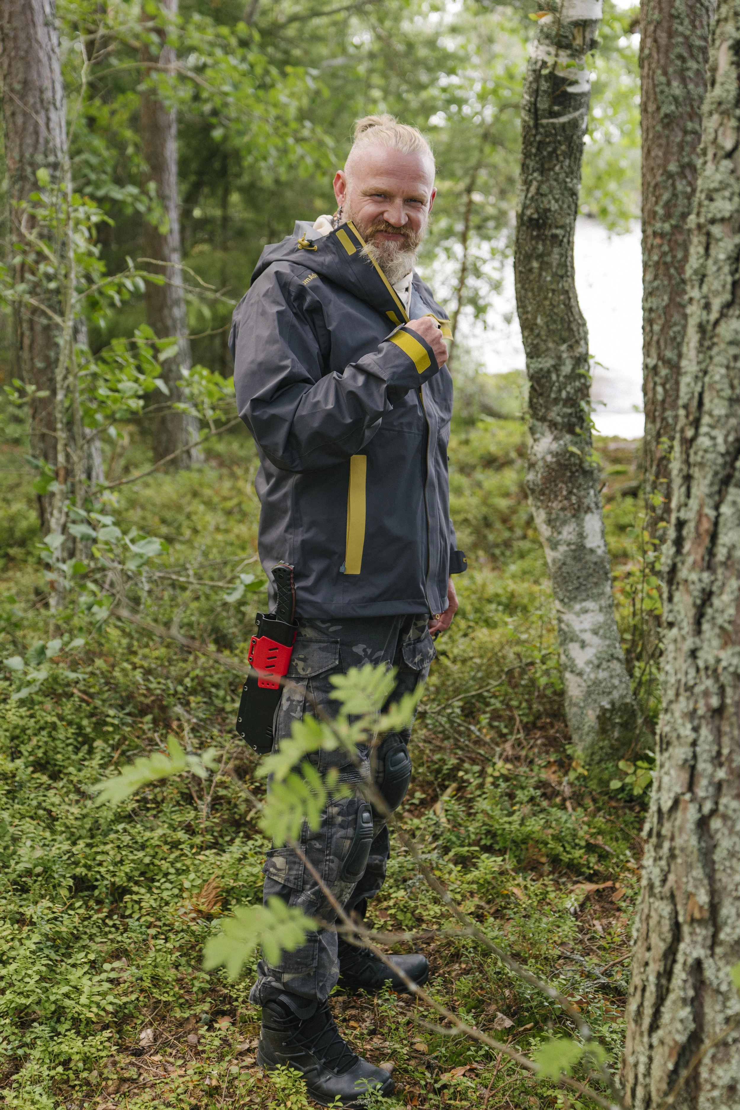 Ein Mann in Outdoor-Kleidung mit Messer an der Hüfte steht in einem Wald und lächelt.