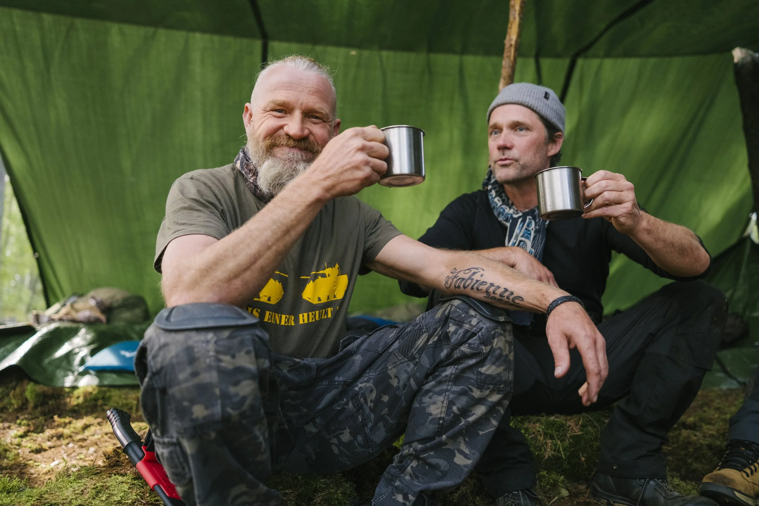 Zwei Männer sitzen in einem Zelt am Boden, trinken aus Metallbechern und genießen eine Pause in der Natur, umgeben von grüner Umgebung und einem Lagerfeuer.