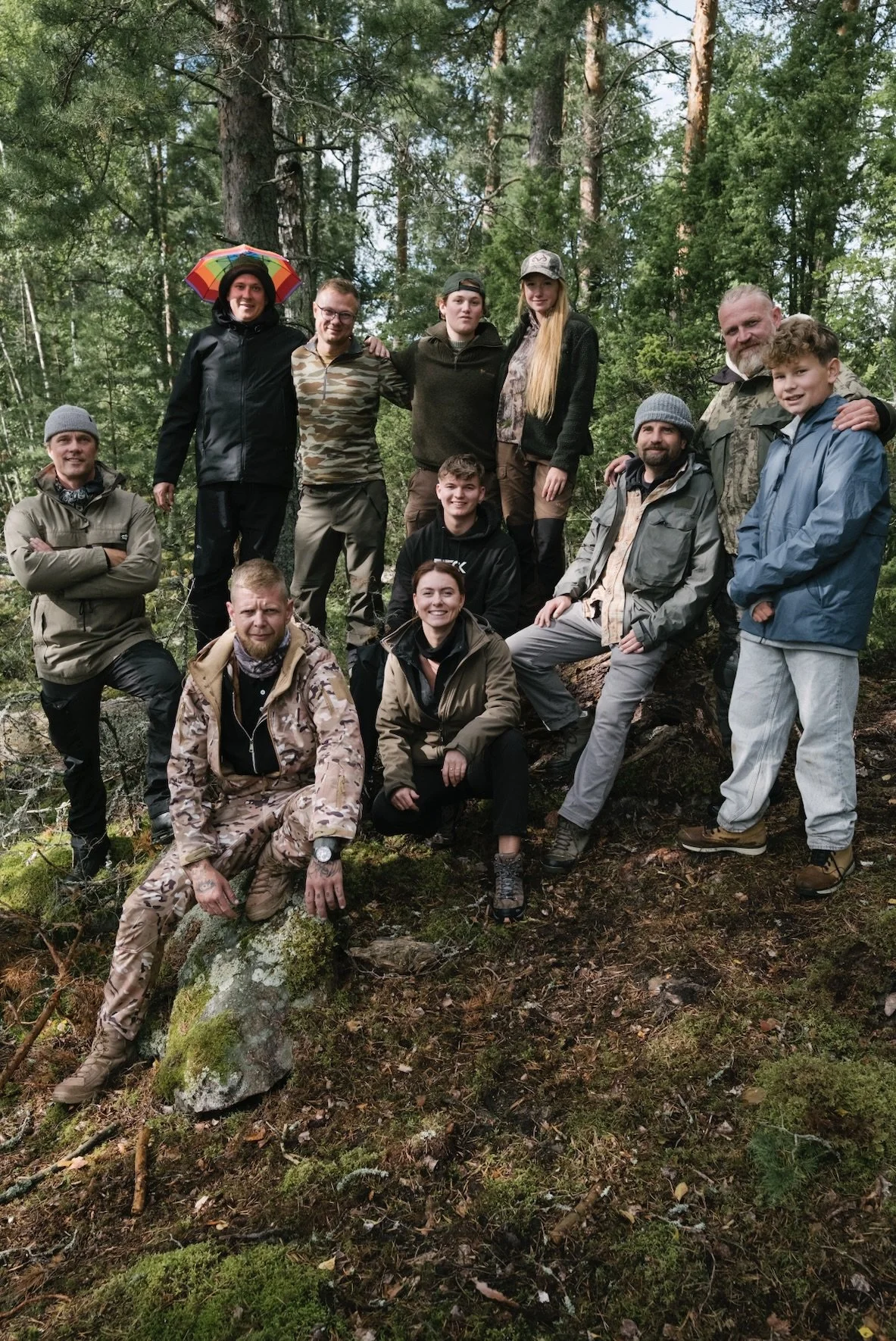 Eine Gruppe von Menschen in einer Waldumgebung, einige sitzen auf einem Baumstamm, andere stehen, alle tragen Outdoor- oder Camouflage-Kleidung.
