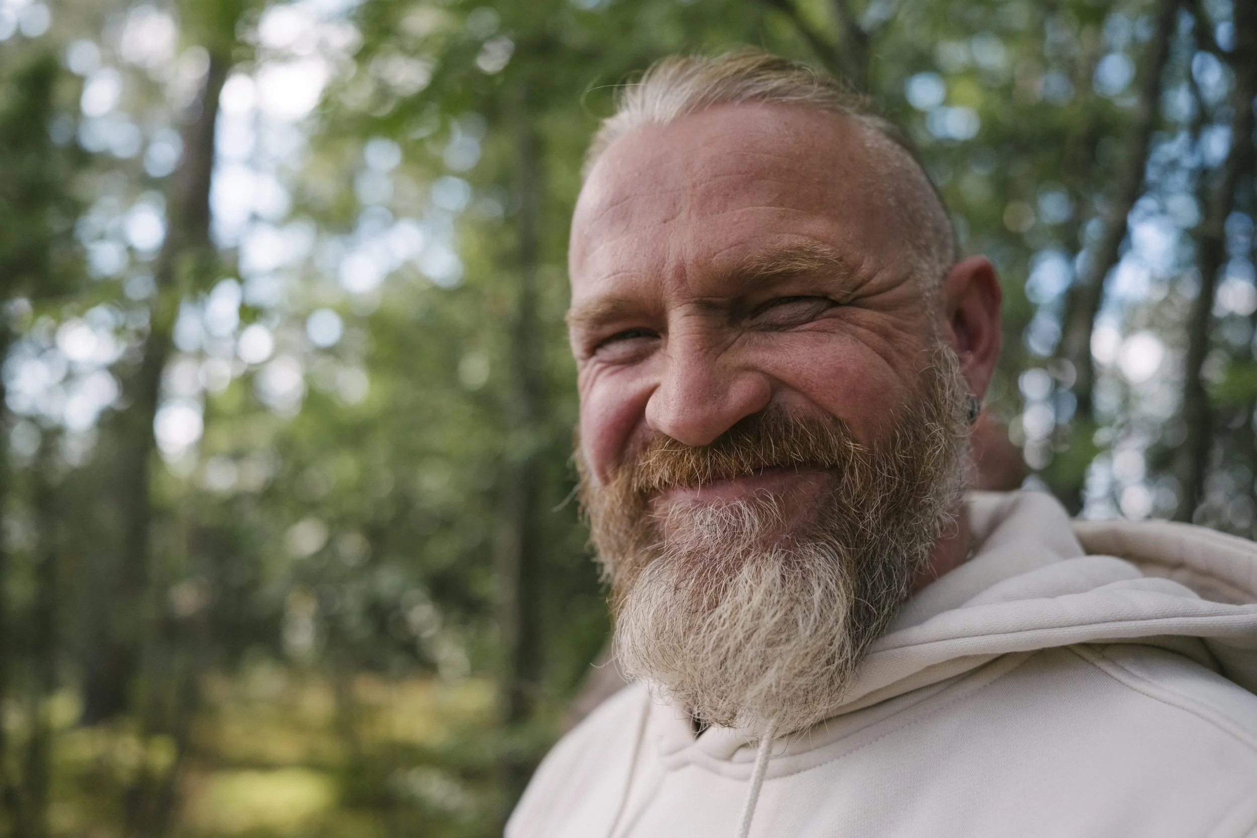 Ein lachender Mann mit Bart in einem Wald bei Sonnenlicht.