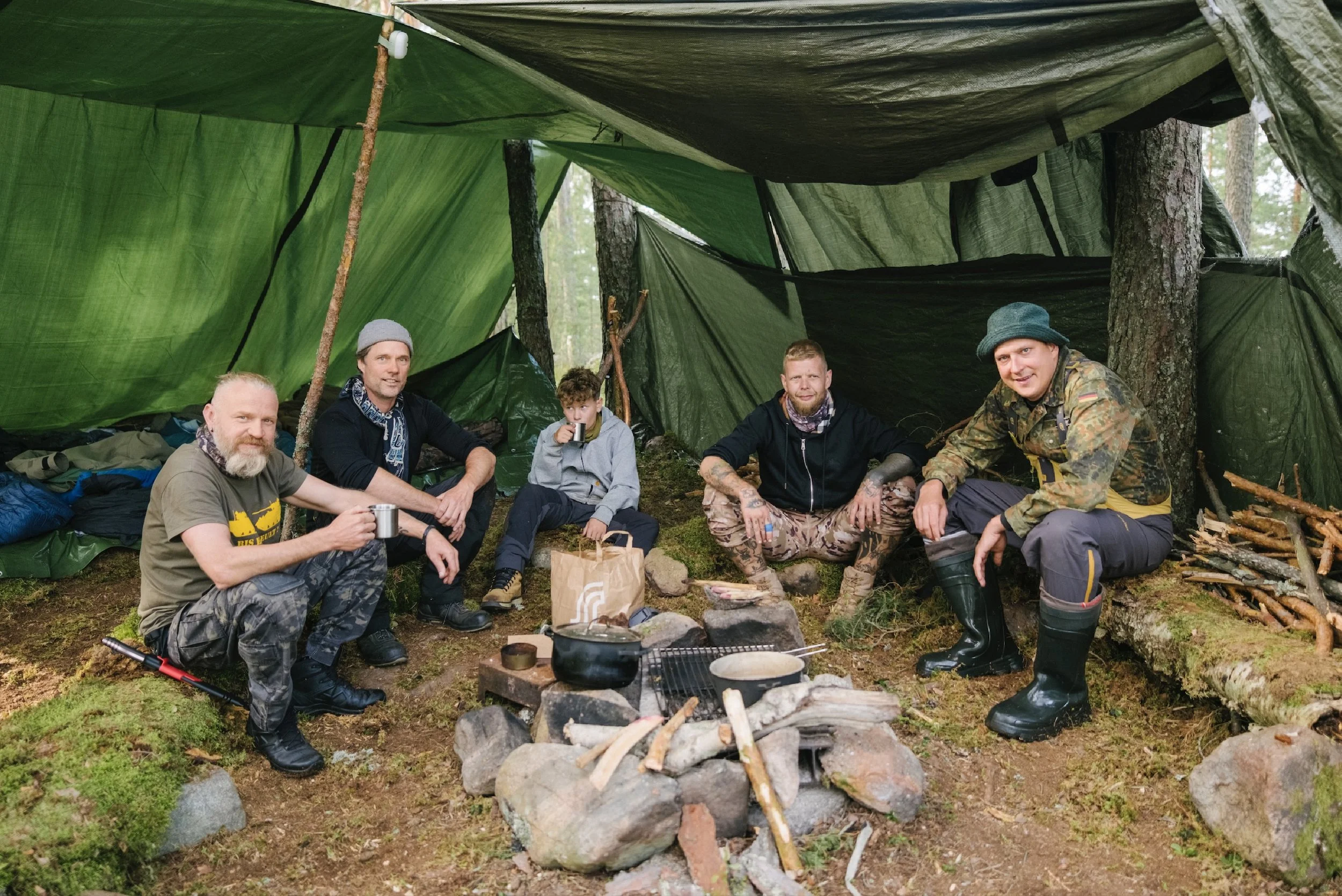Fünf Männer und ein Junge sitzen in einem Zelt im Wald, sie essen und trinken, umgeben von Campingausrüstung und Kochausrüstung