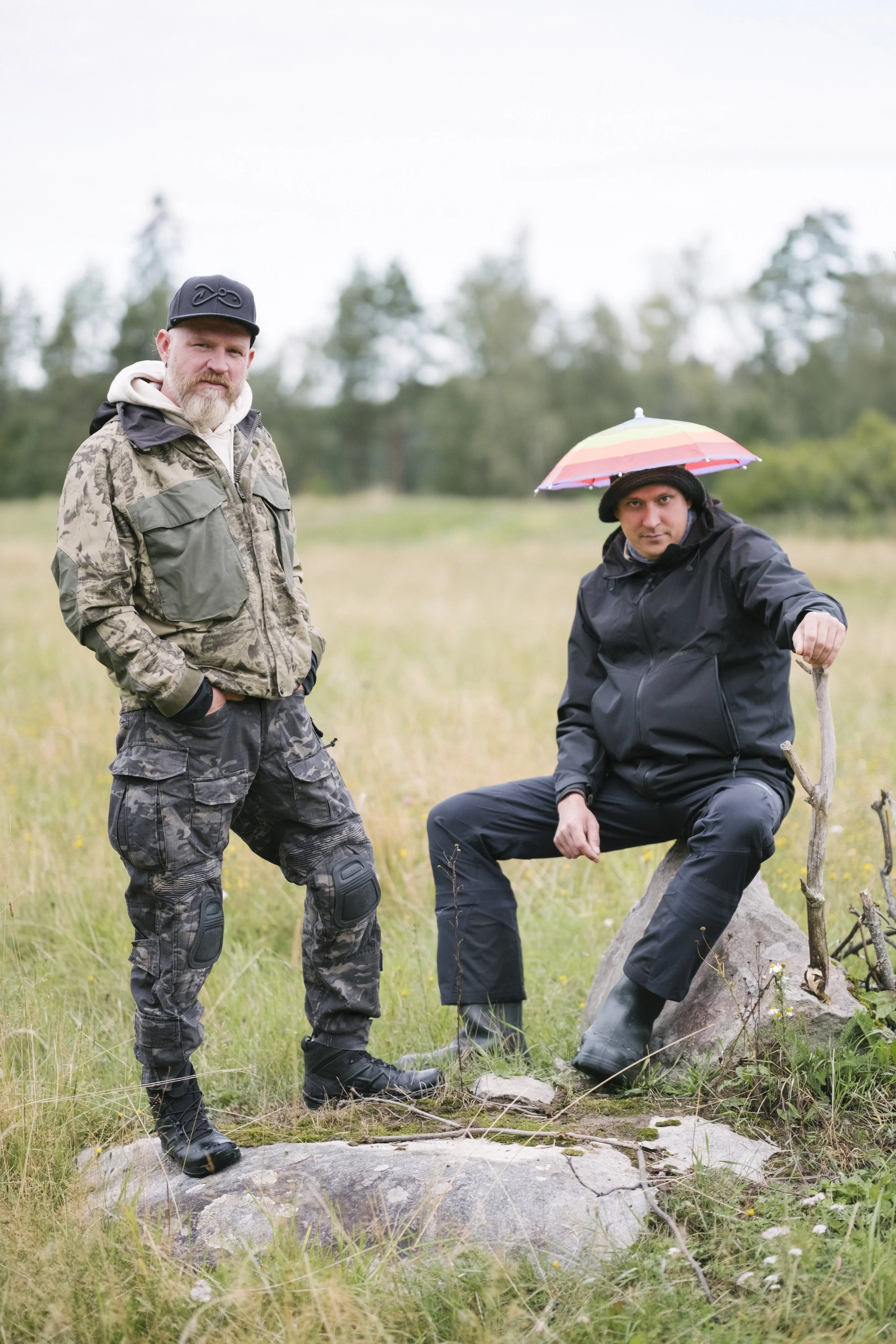 Zwei Männer in Outdoor-Kleidung stehen in einer Wiese, einer sitzt auf einem Stein mit einem Regenschirm auf dem Kopf, der andere steht daneben.