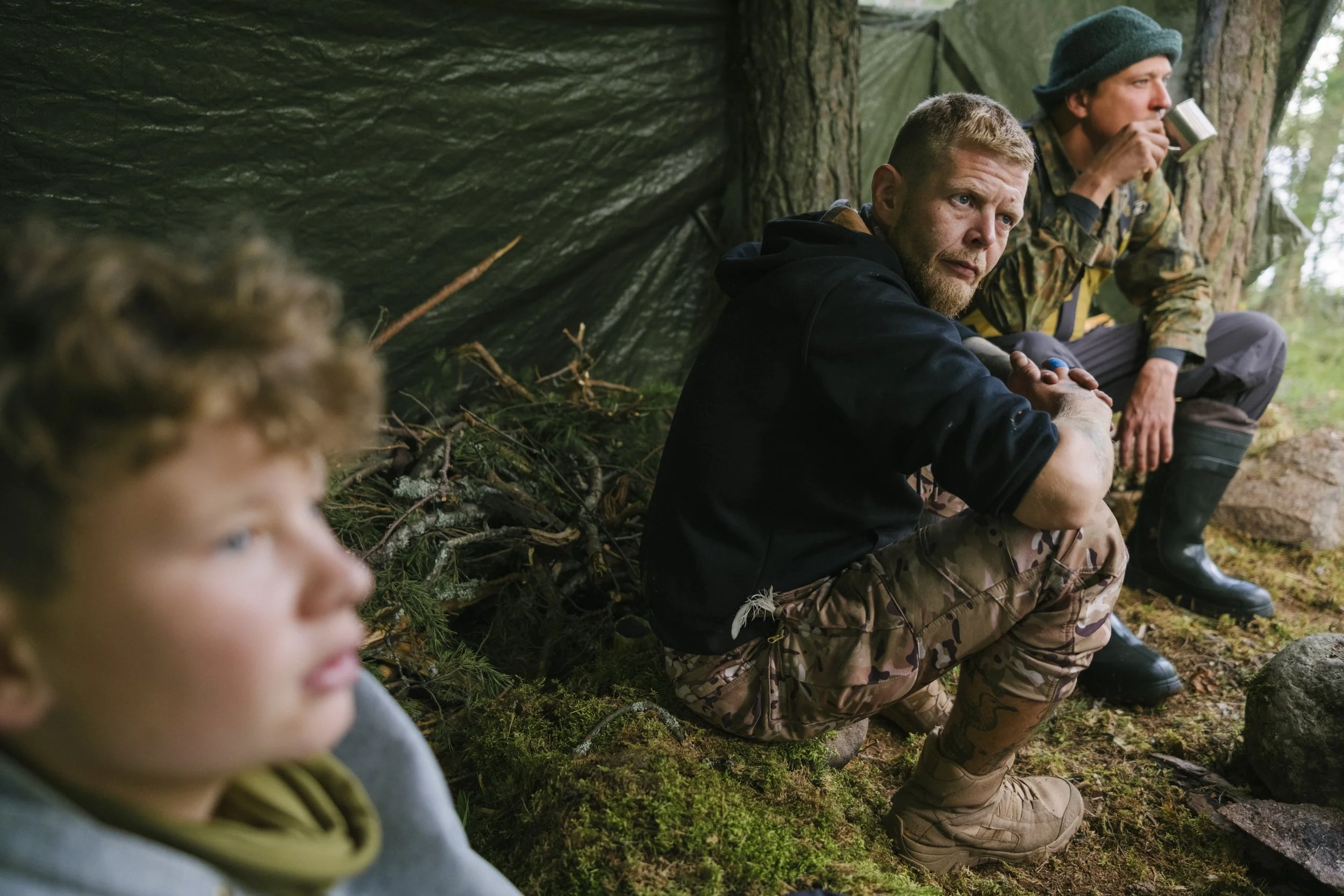 Drei Männer und ein Junge sitzen im Wald, einer trägt Camouflage, alle trinken Getränke und sitzen vor einer Tarnhütte.