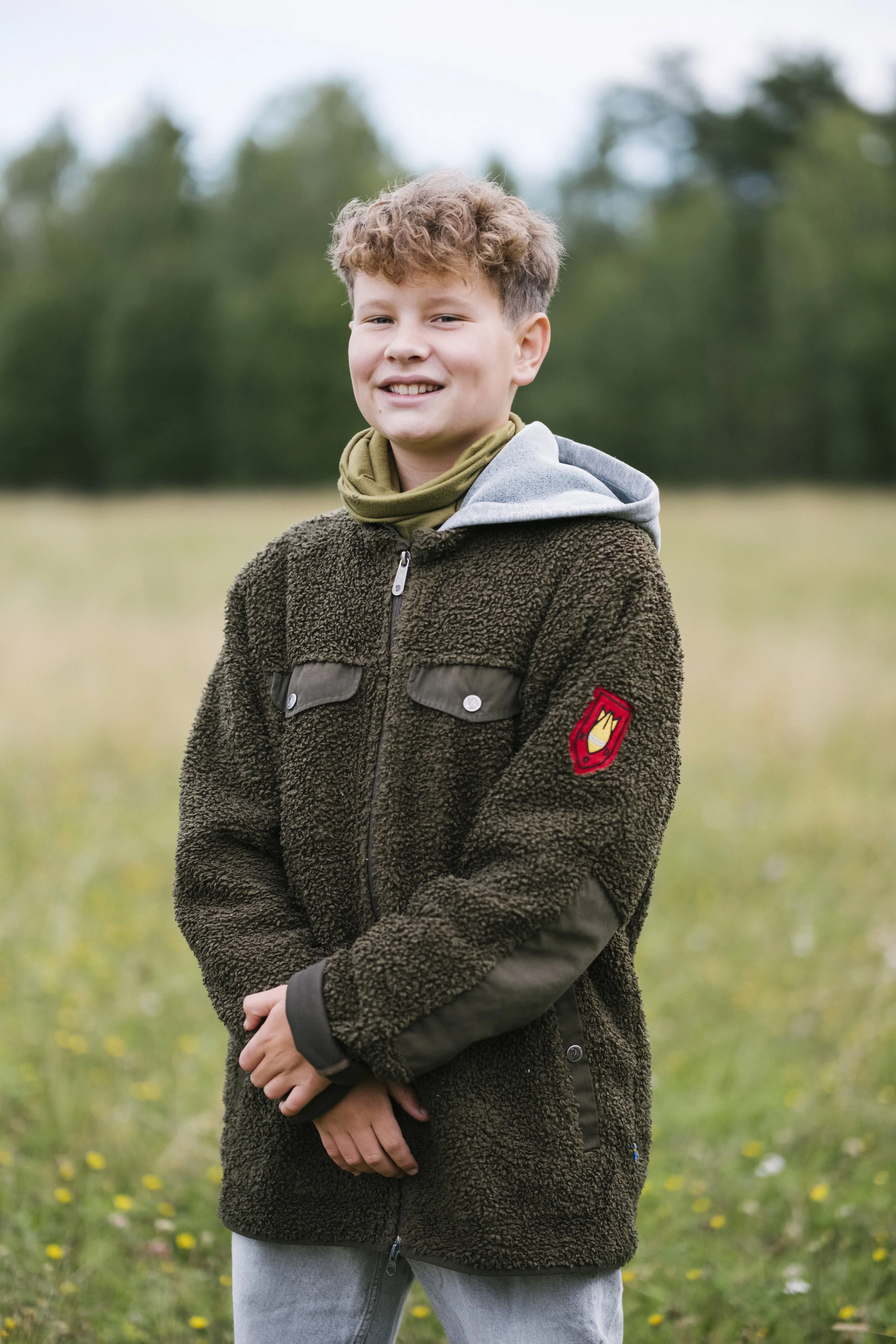 Ein lachender Junge in einem Wald im Freien trägt eine braune Fleece-Jacke, eine graue Kapuzenjacke und einen grünen Schal.