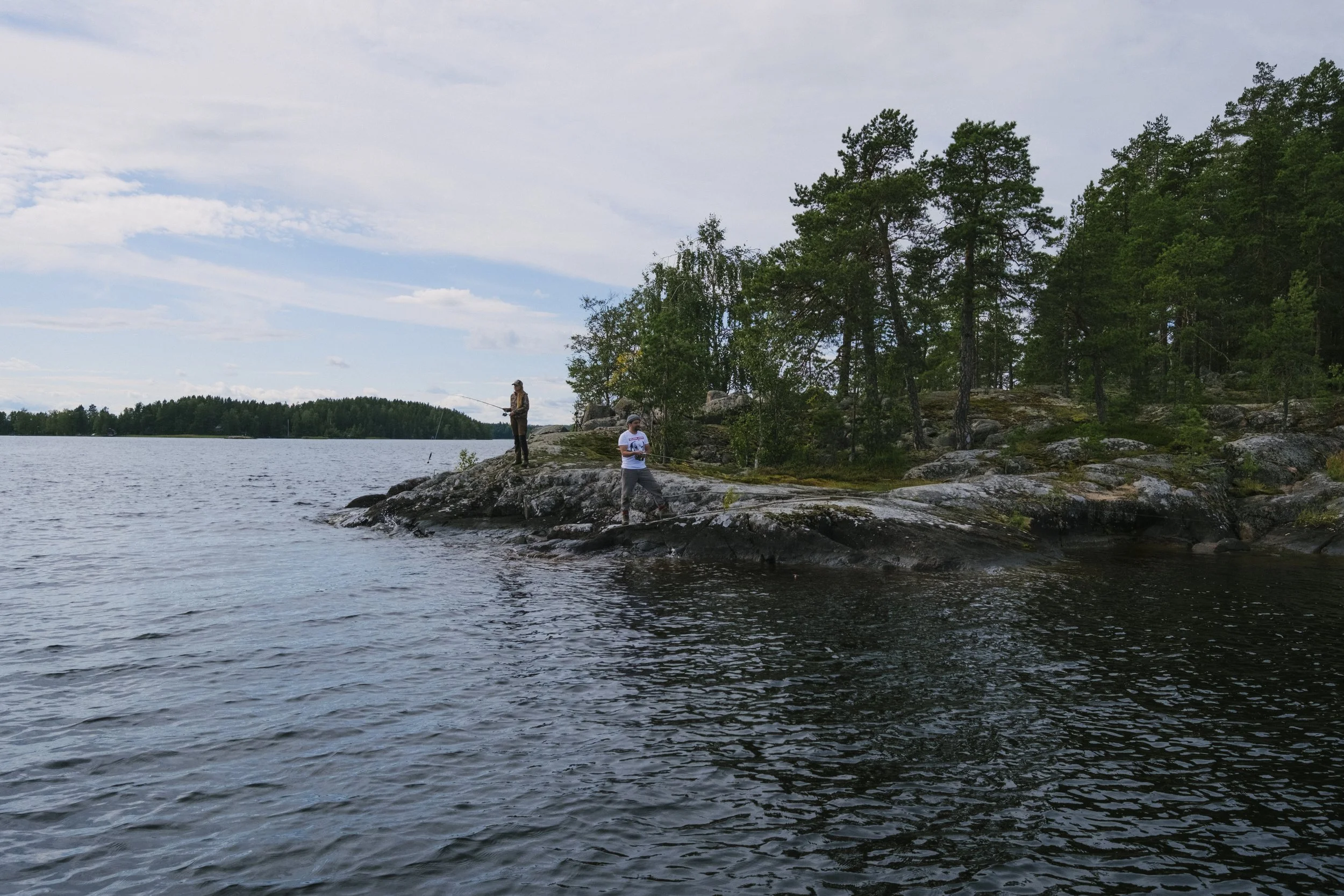 Zwei Menschen stehen auf einer felsigen Küste an einem See, einer fischt, während der andere mit einem Tablet oder Smartphone vor dem Wasser steht, umgeben von Bäumen und einem bewölkten Himmel.