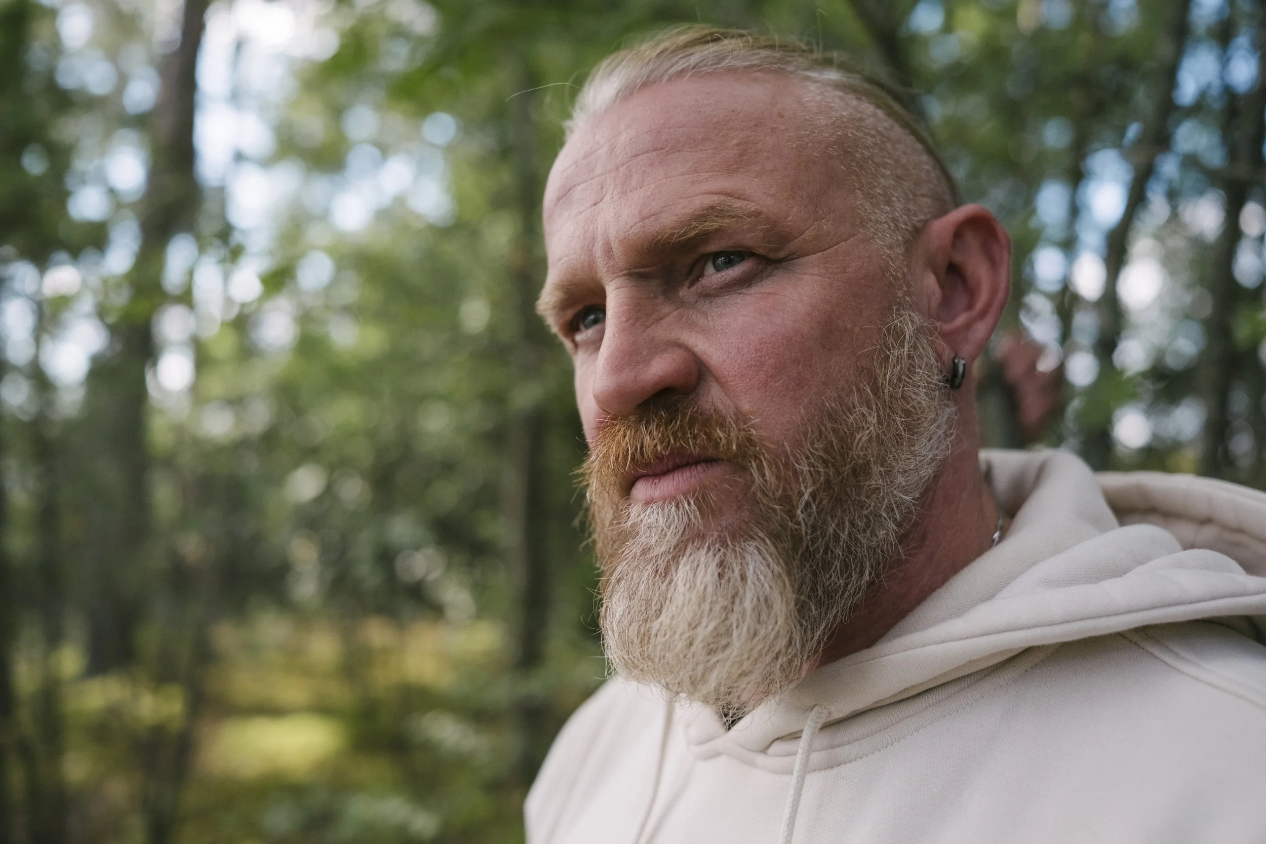 Nahaufnahme eines Mannes mit blondem Bart, blondem Haarschnitt und Ohrring in der rechten Ohrmuschel im Wald