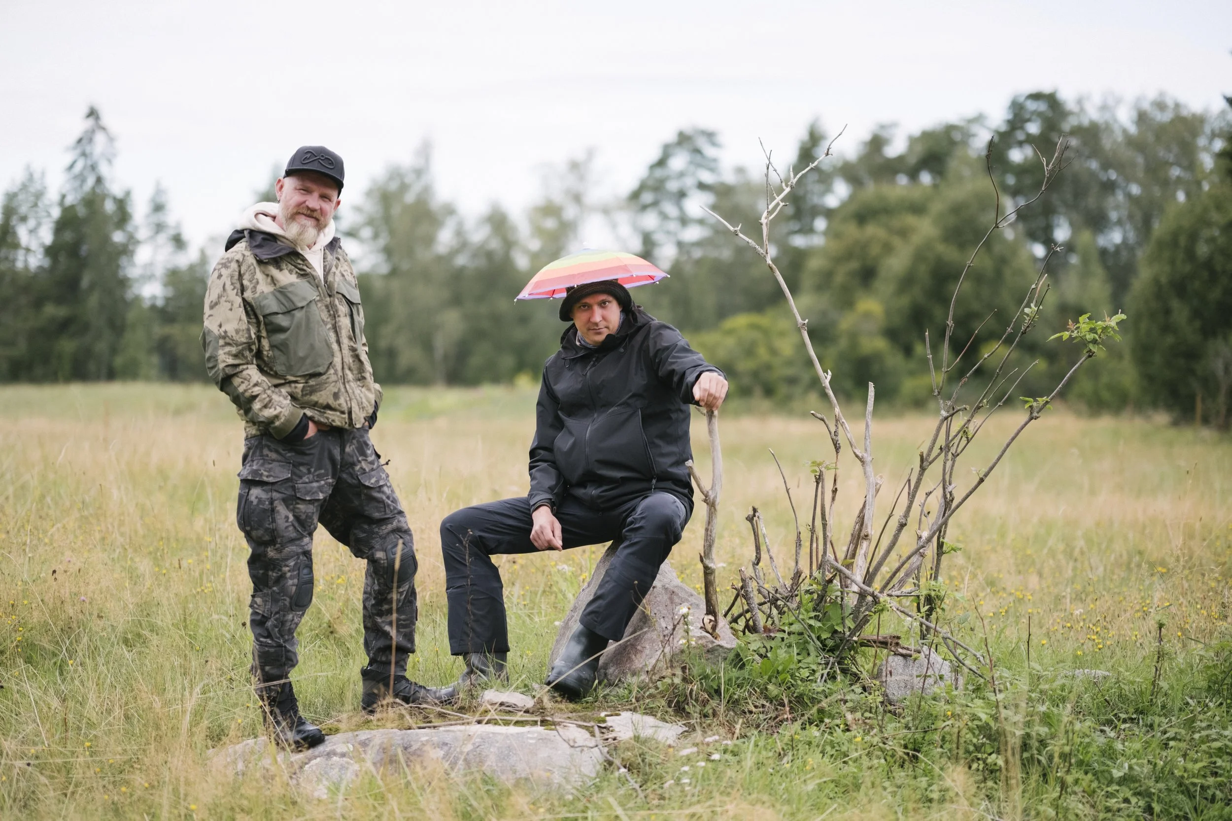 Zwei Männer stehen in einem Feld. Einer sitzt auf einem Baumstumpf mit einem roten Regenschirm, der andere steht neben ihm. Beide tragen dunkle Outdoor-Kleidung, während es leicht regnet.