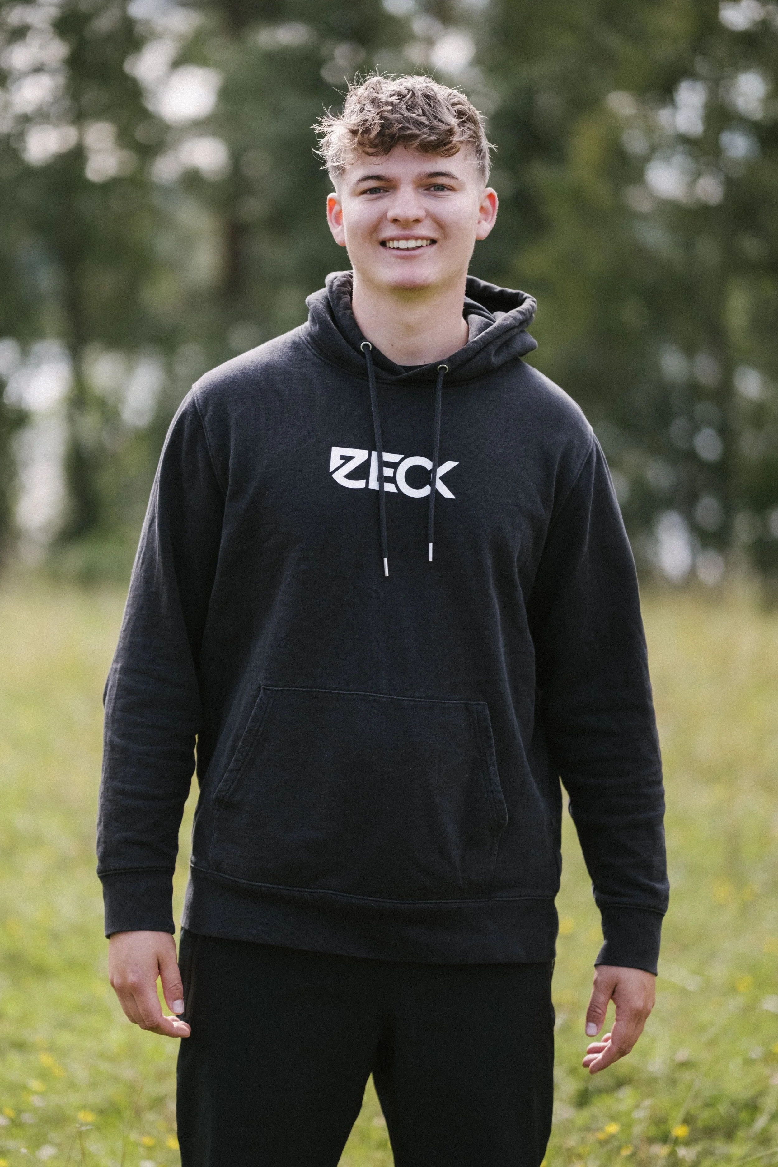 Junger Mann trägt eine schwarze Hoodie mit weißem Schriftzug, steht im Freien vor grüner, unscharfer Natur.