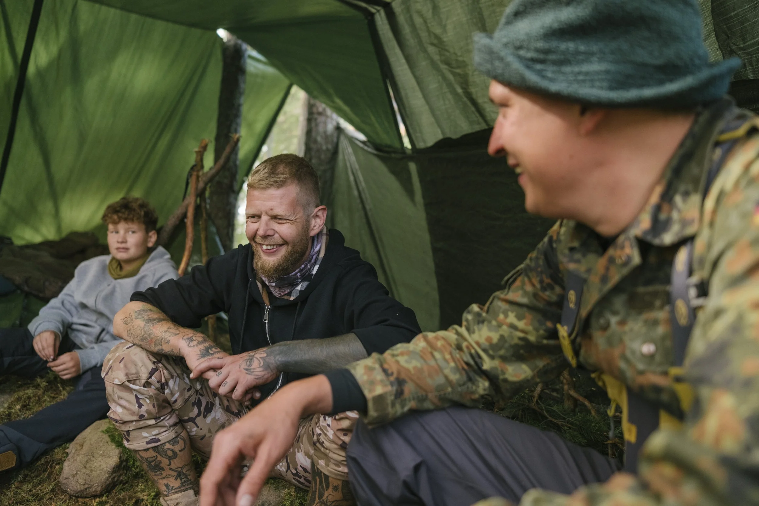Drei Leute sitzen in einem grünen Zelt im Wald, lachen und unterhalten sich.