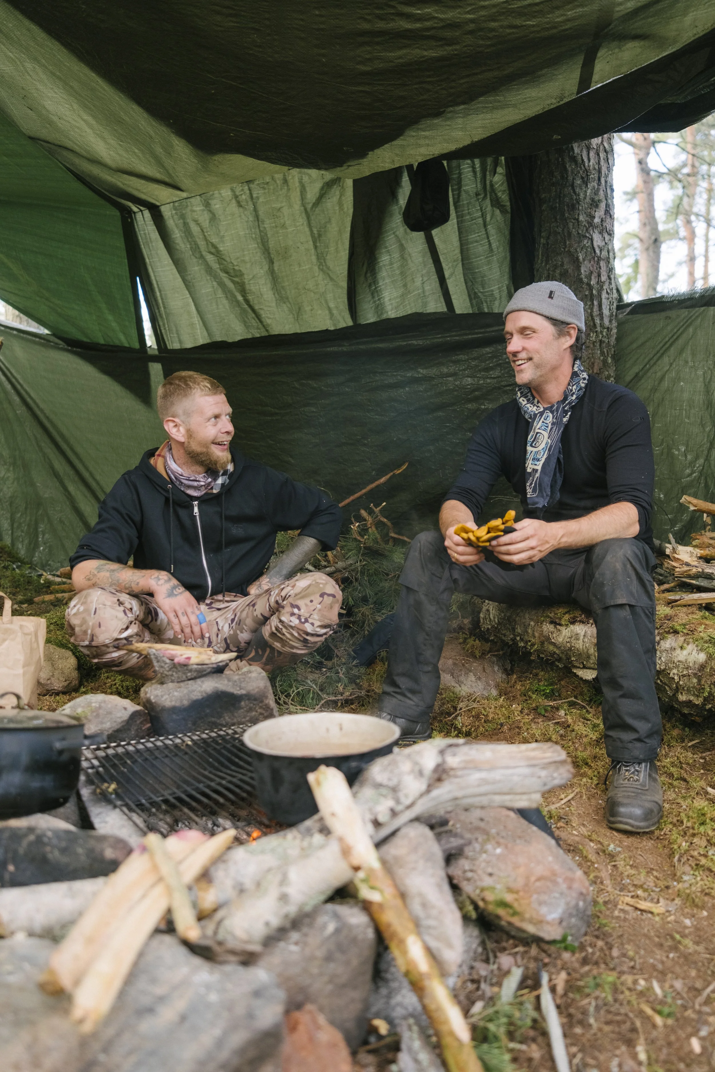 Zwei Männer sitzen in einem Zelthering im Wald, unter einem Notdach, die eine Mahlzeit zubereiten und sich unterhalten.