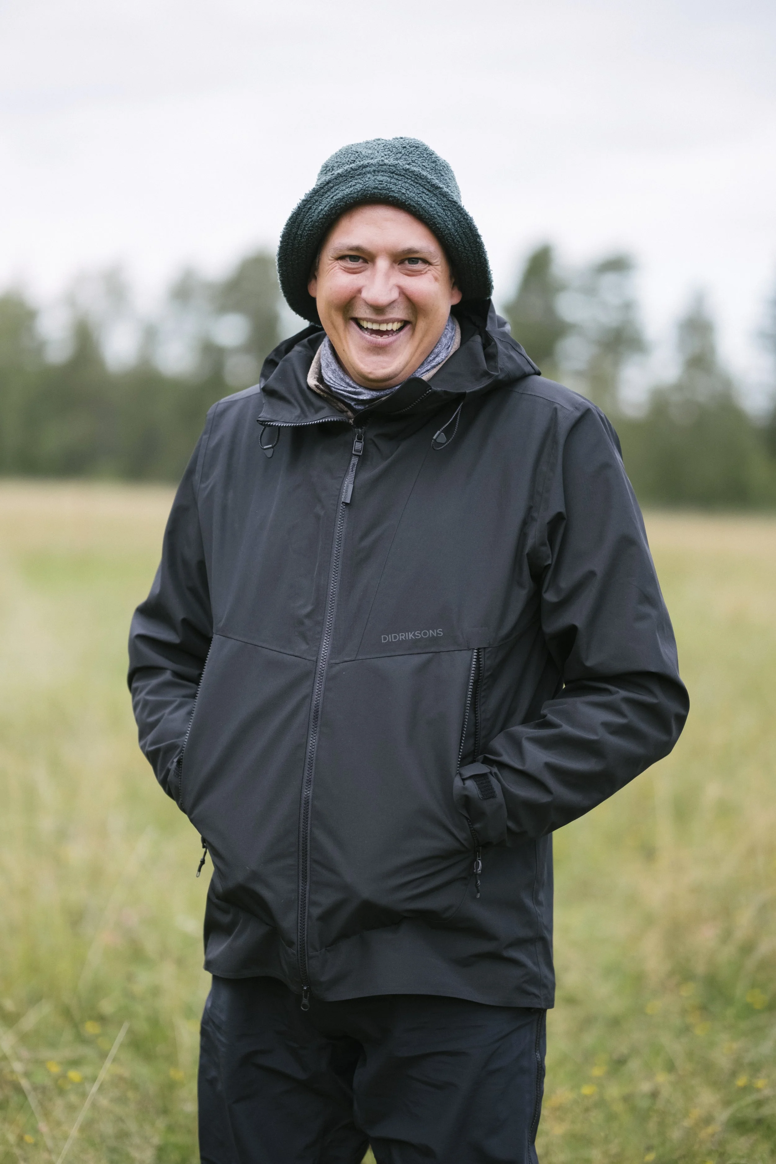 Ein lachender Mann trägt eine schwarze Jacke und einen Wollmütze, steht in einer Wiese mit Bäumen im Hintergrund.