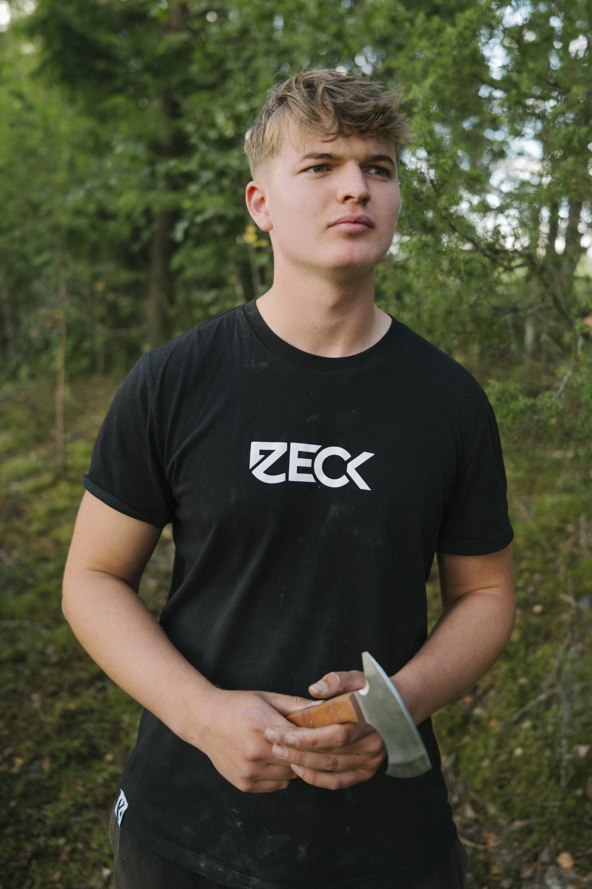 Junge Mann im schwarzen T-Shirt mit dem Schriftzug "ZECK" steht im Wald und hält ein Messer in der Hand.