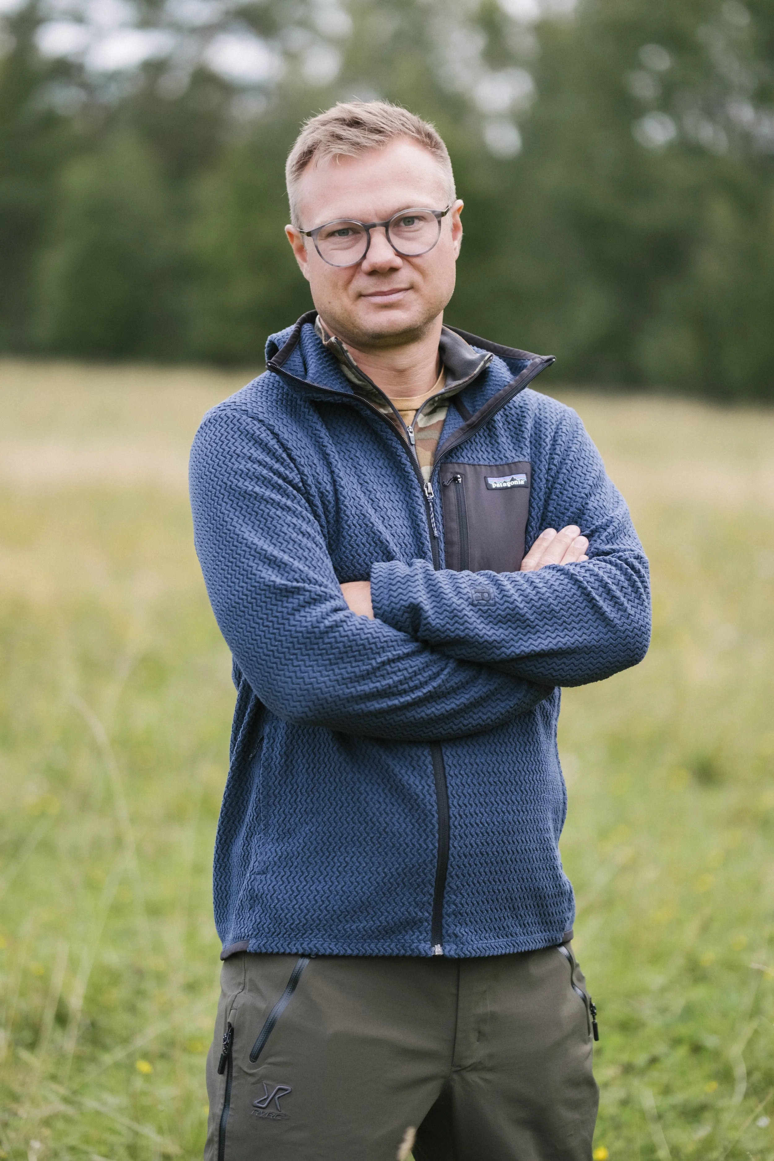 Mann in Outdoors-Umgebung, trägt eine blaue Jacke und braune Hosen, steht mit verschränkten Armen, im Hintergrund Bäume und Wiesen.
