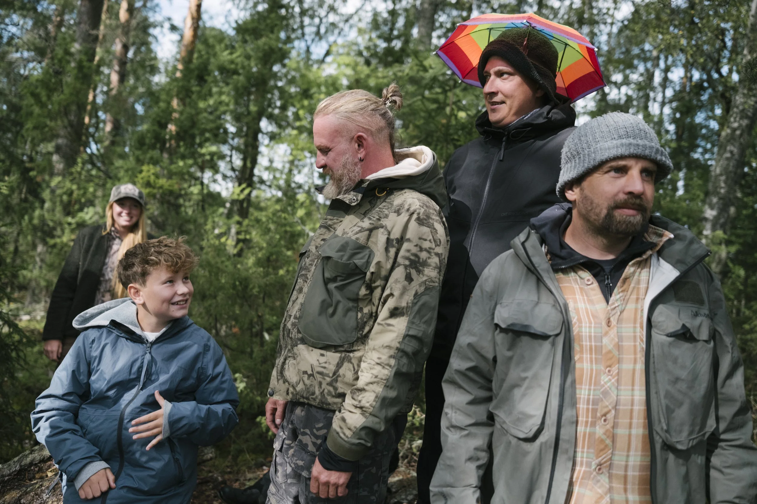 Fünf Menschen in einem Wald, lächelnd und im Gespräch, dekoriert mit Regenbekleidung und einem bunten Regenschirm, beim Wandern.