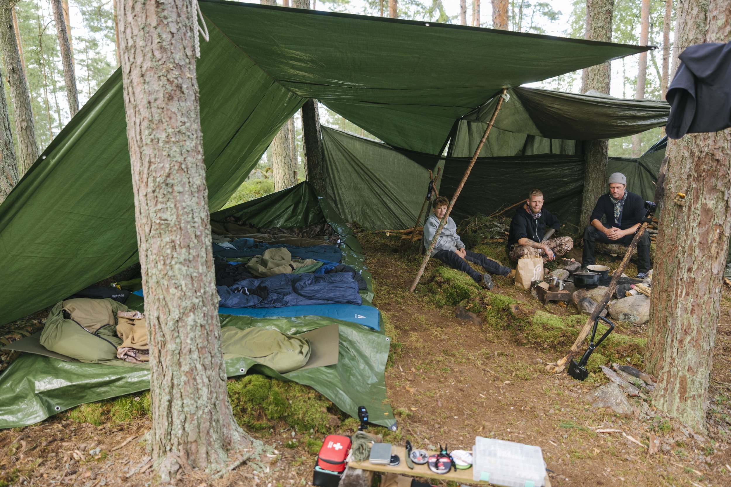Drei Personen sitzen in einem Zeltdorf im Wald um ein Lagerfeuer. Es gibt mehrere Schlafsäcke und Campingausrüstung auf dem Boden.