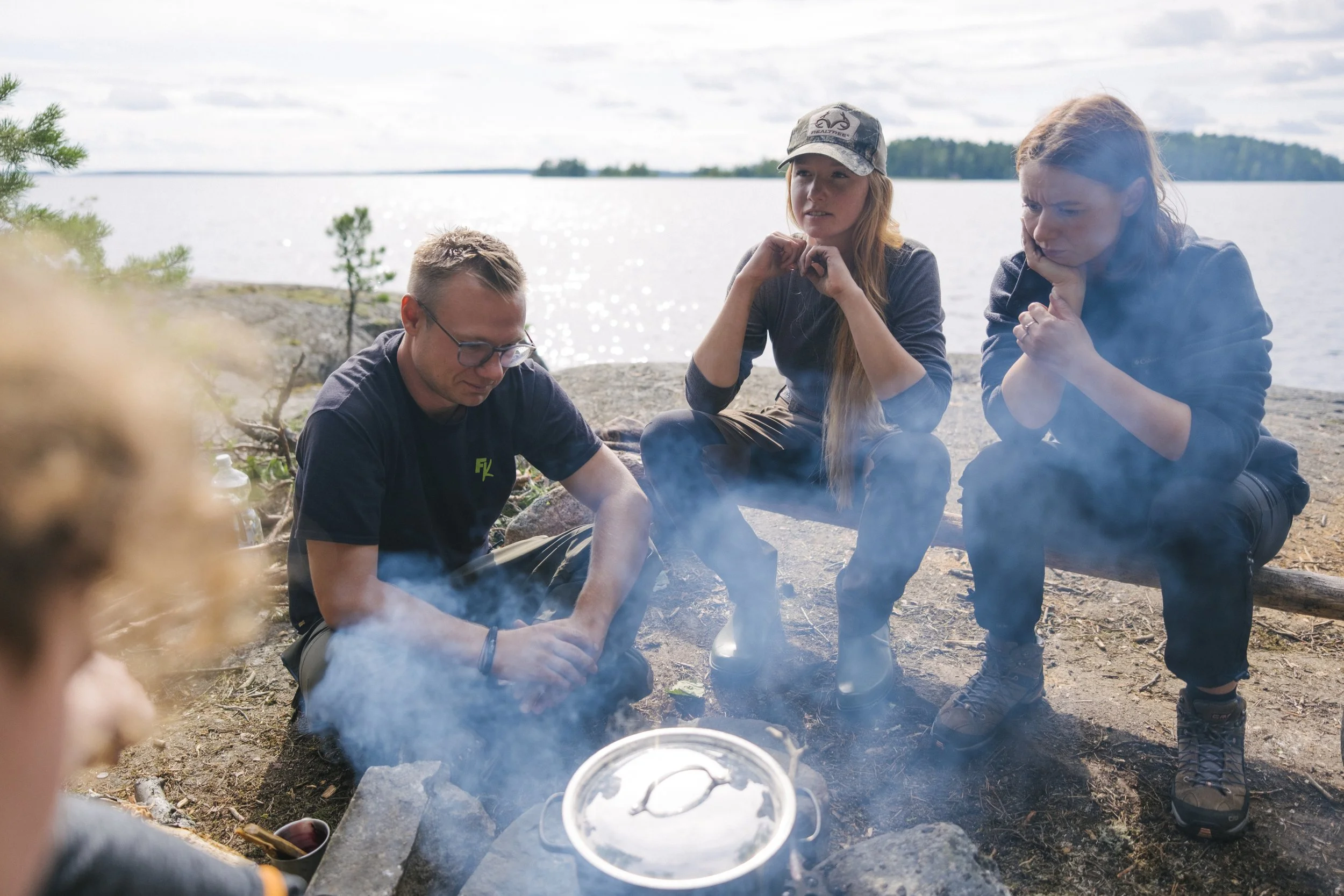 Drei Menschen sitzen am Lagerfeuer am Wasser, während sie sprechen und nachdenken, umgeben von Natur mit Wasser und Bäumen im Hintergrund.