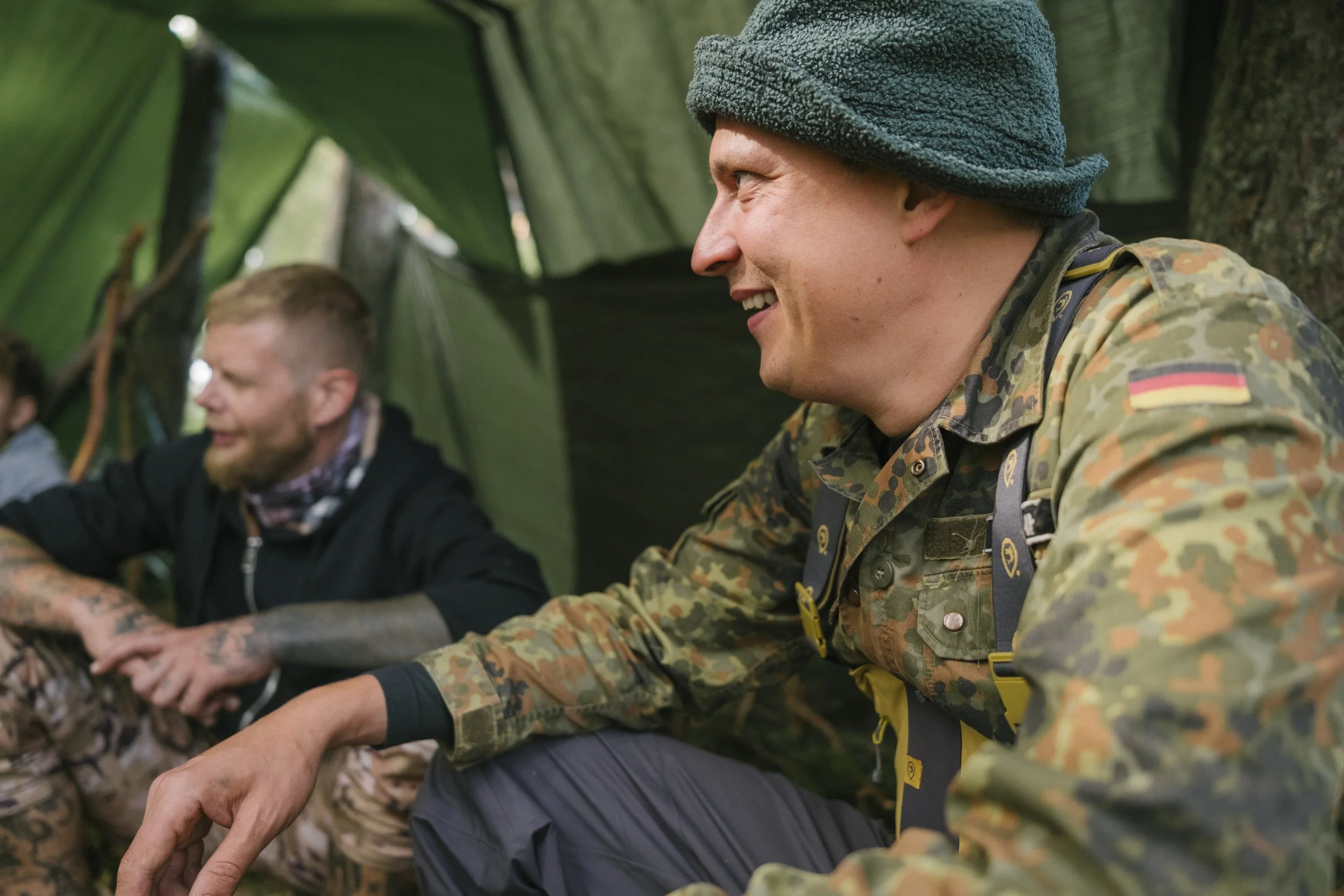 Zwei Männer sitzen im Zelt, lachen und unterhalten sich. Einer trägt Camouflage-Kleidung mit deutscher Flagge, der andere trägt dunkle Kleidung. Die Stimmung ist fröhlich.