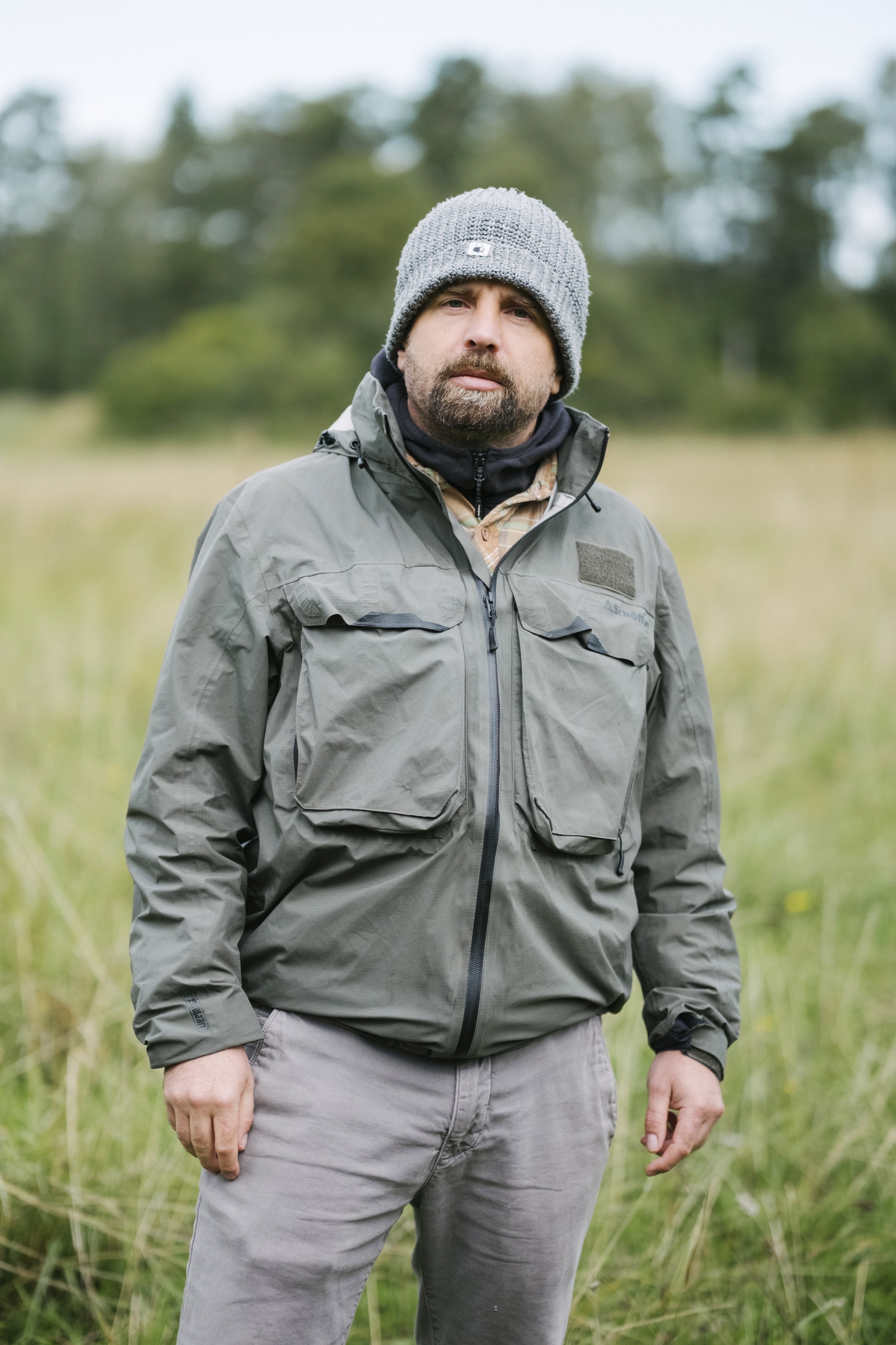 Mann trägt eine graue Jacke und Mütze in einem Feld mit Bäumen im Hintergrund.