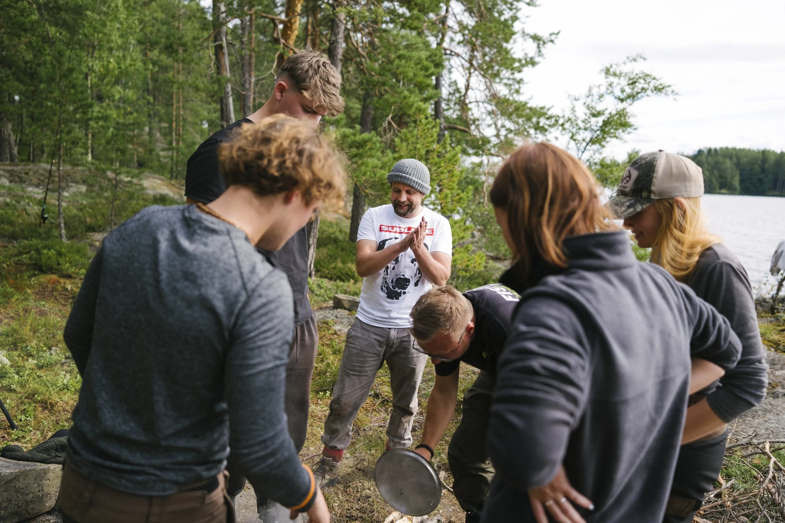 Gruppe von sechs jungen Leuten im Freien am See, die einen Gegenstand untersuchen, während ein Mann im weißen T-Shirt und grauem Hut daneben steht und sich freut.