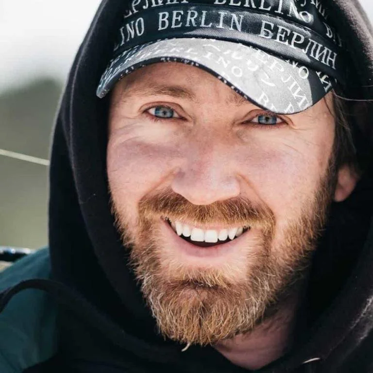 Lächelnder Mann mit blauäugigen, rötlichen Haaren und Bart, trägt eine schwarze Kappe mit weißen Aufschriften.