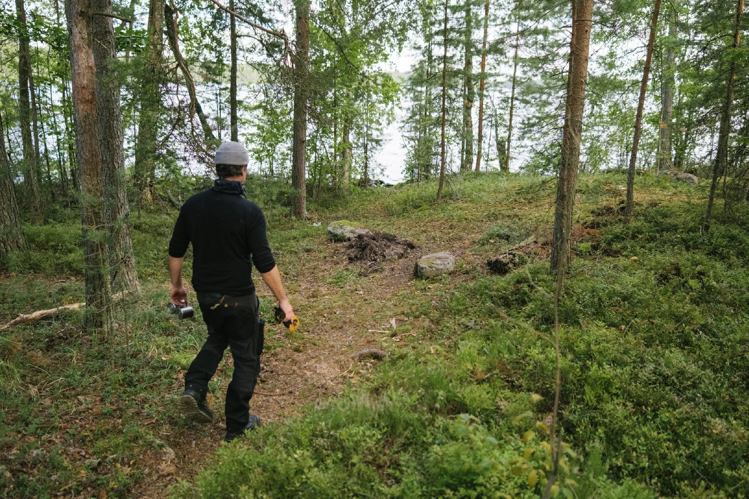 Ein Mann in dunkler Kleidung und grauem Hut wandert durch einen grünen, bewaldeten Pfad in der Nähe eines Sees oder Flusses