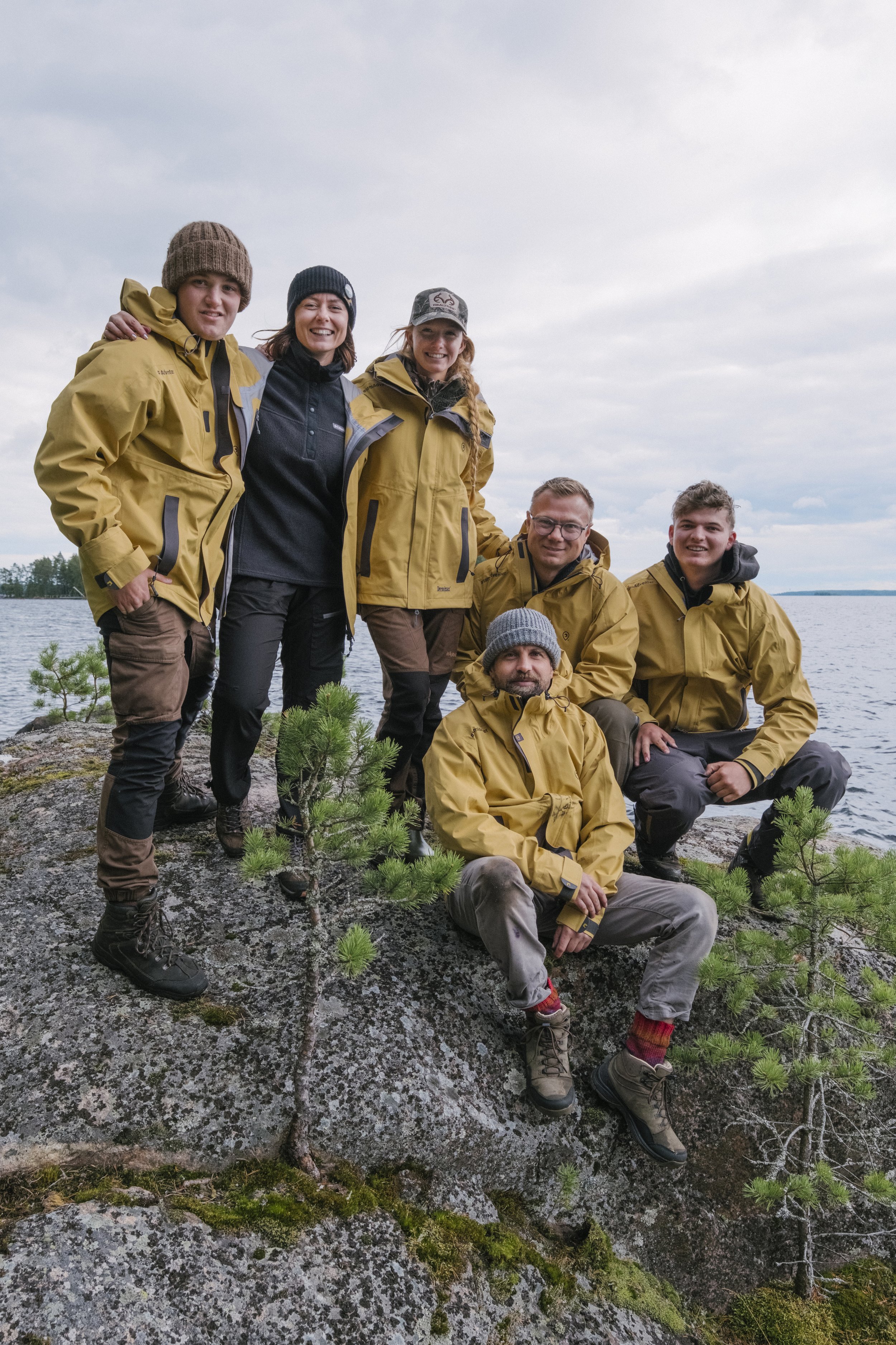 Gruppe von sieben Menschen in Outdoor-Kleidung steht und sitzt auf einem Felsen am Ufer eines Sees, umgeben von kleinen grünen Bäumen bei bewölktem Himmel.