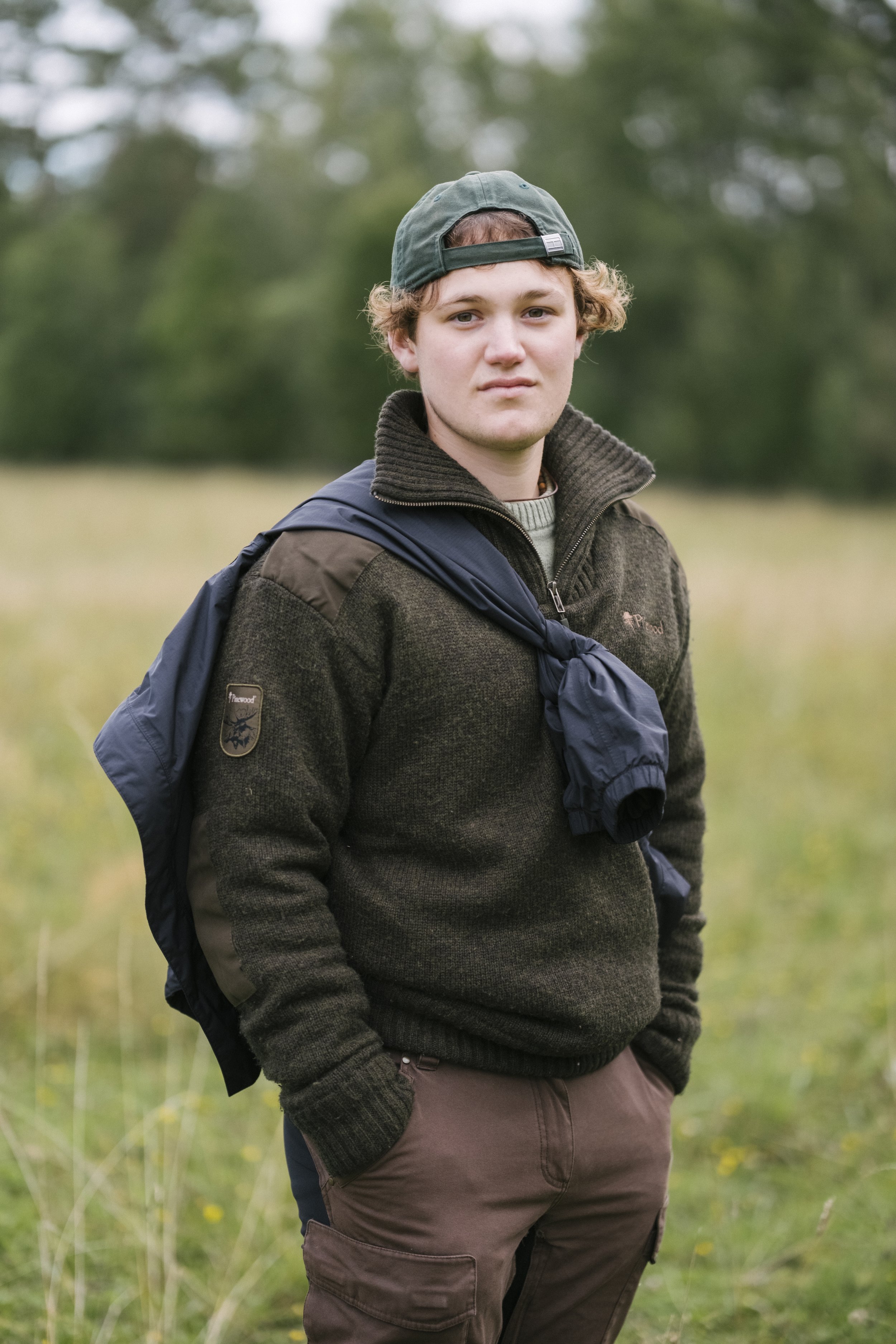 Ein junger Mann steht im Freien in einer grünen Wiese mit Bäumen im Hintergrund, trägt eine grüne Basecap, eine braune Jacke mit einer weiteren Jacke darüber, die um den Hals gebunden ist, und braune Hosen.