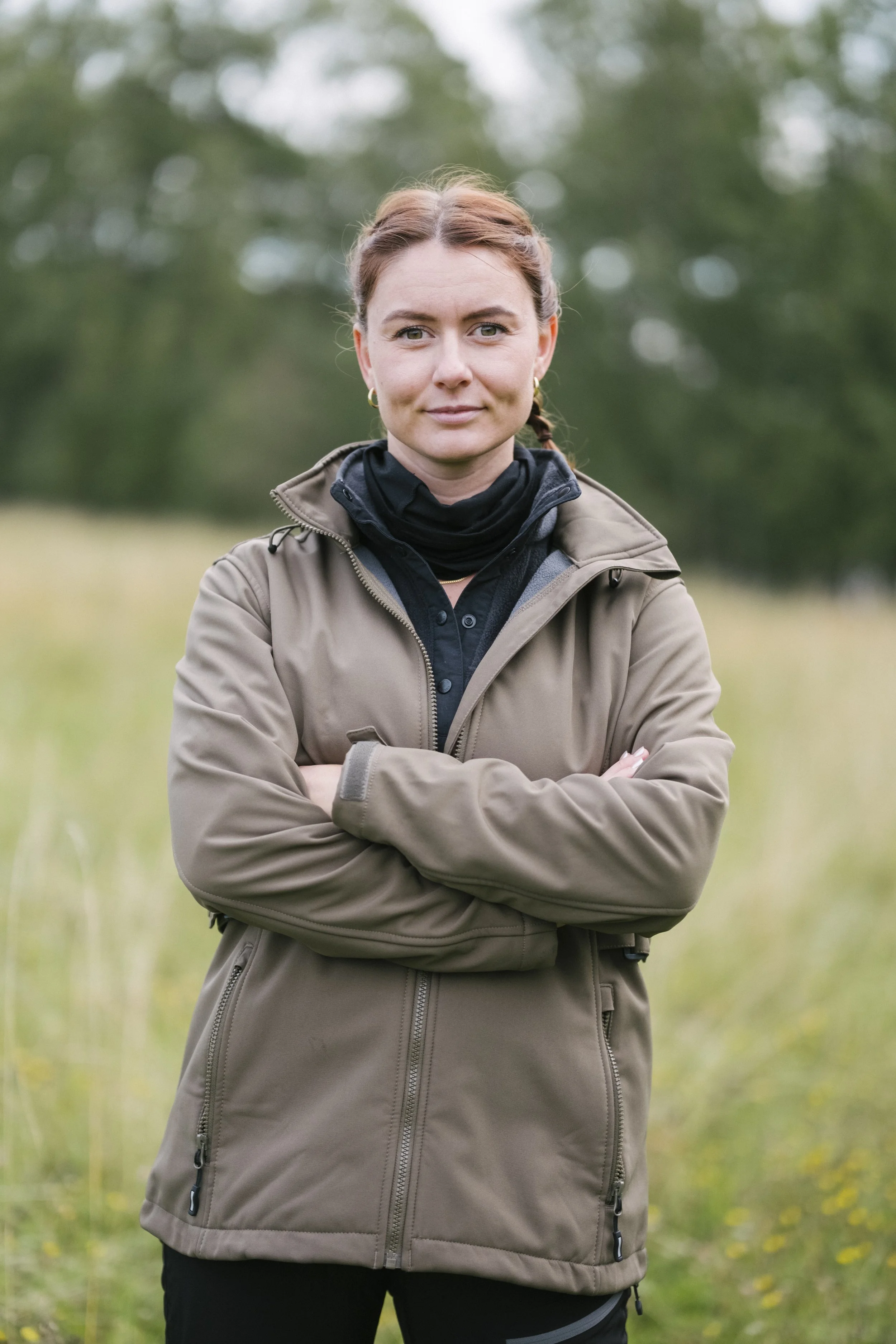 Eine Frau steht im Freien mit verschränkten Armen, trägt eine beige Jacke und schaut in die Kamera, im Hintergrund sind grüne Bäume und eine Wiese.