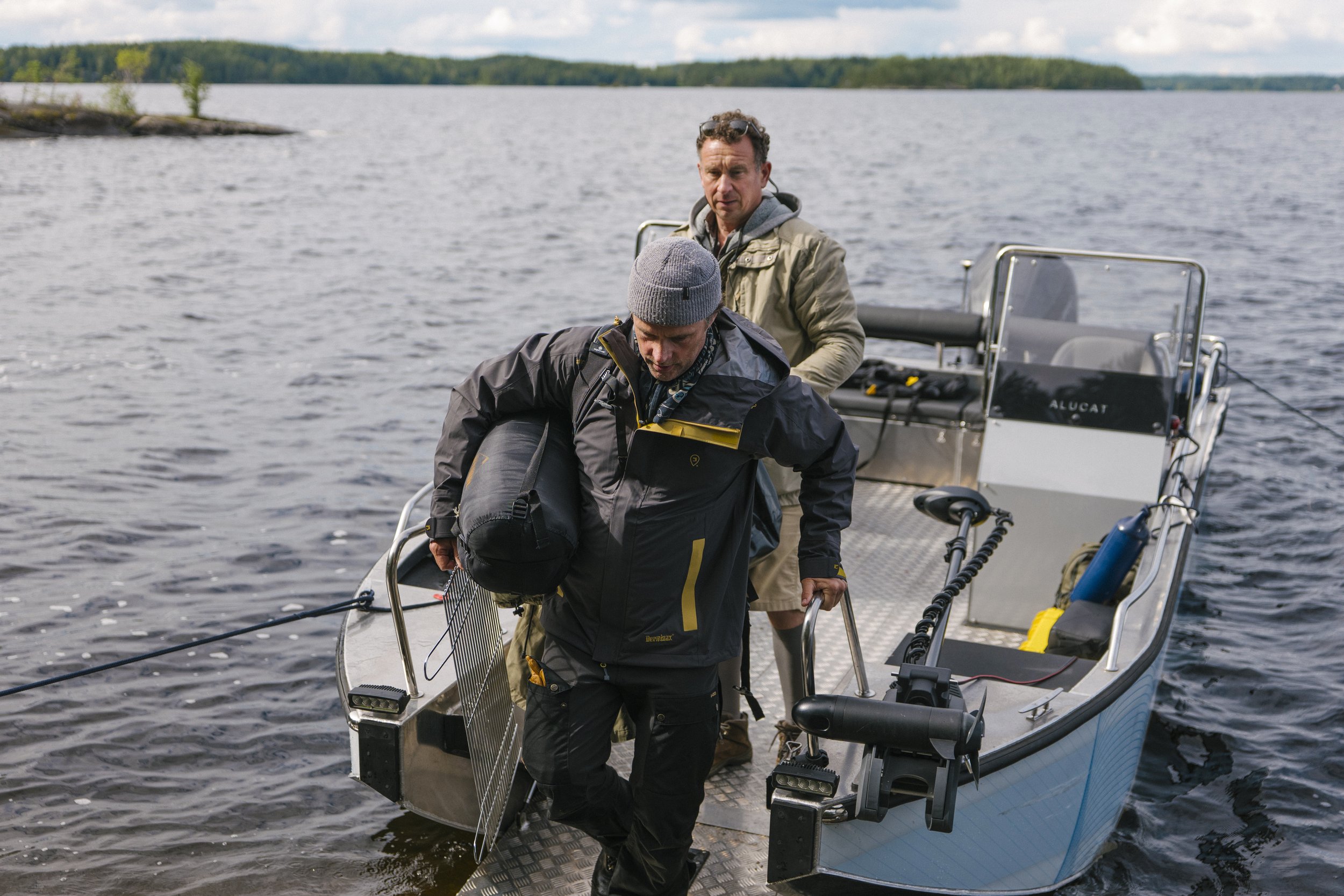 Zwei Männer steigen in ein kleines Motorboot auf einem Seenufer, einer trägt eine schwarze Jacke und einen grauen Wollmützen, der andere steht hinter ihm, beide sind bereit für eine Bootstour.