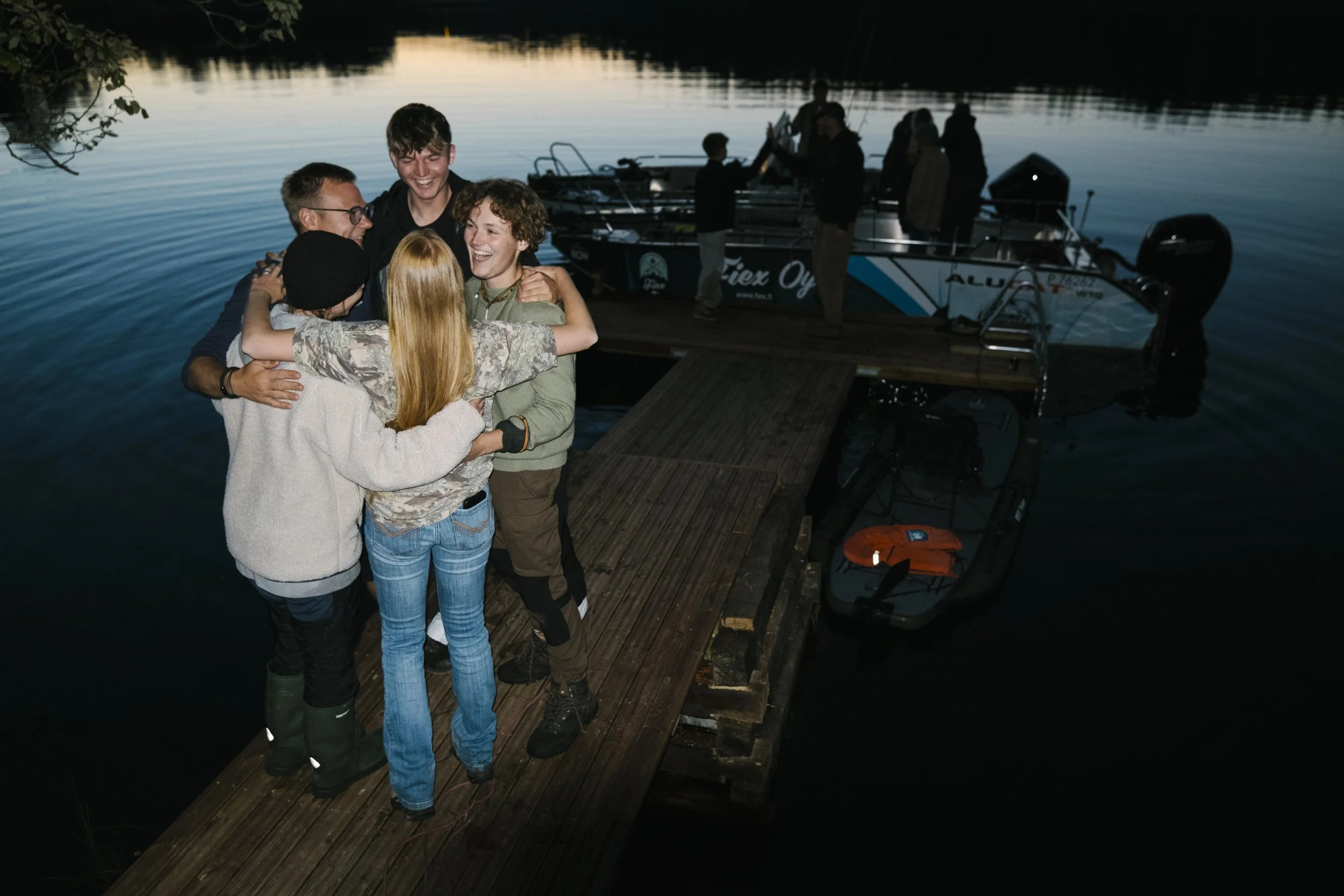 Gruppe von Freunden, die sich umarmen und lachen auf einem Steg am See bei Sonnenuntergang, Boote im Wasser