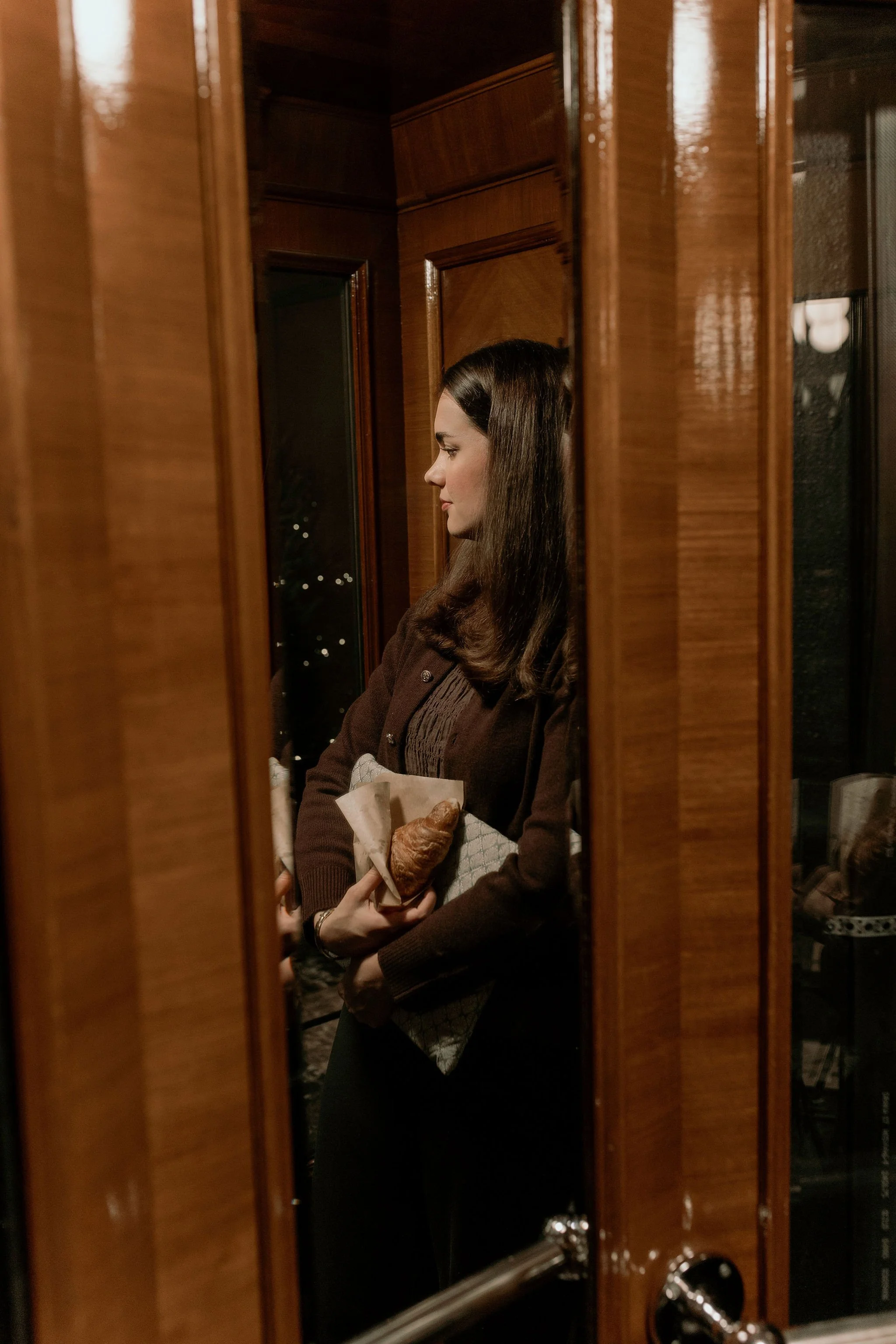 A woman with brown hair is seen through a wooden door lock with glass, holding a breakfast pastry and a wrapped item, standing inside near a window.
